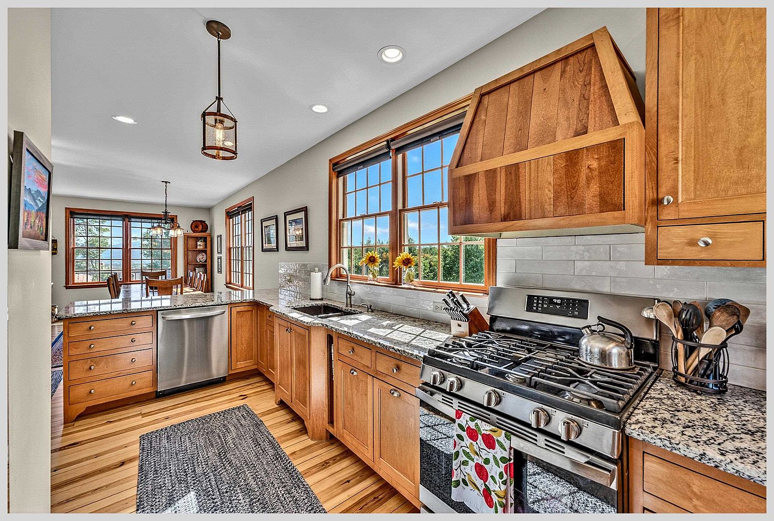 This warm and inviting kitchen features light wood cabinetry, granite countertops, and stainless steel appliances, including a gas range with a custom wood vent hood. A large window above the sink provides ample natural light and a view of the outdoors, while a patterned runner rug adds texture to the wood flooring. The space flows seamlessly into an adjacent dining area, creating an open and functional layout perfect for daily living.