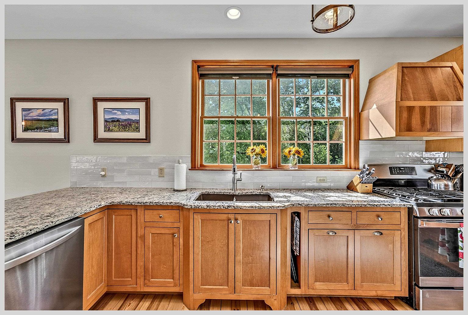 This kitchen features warm-toned wood cabinetry paired with speckled stone countertops and a light-colored subway tile backsplash. A central sink is positioned beneath a large, multi-pane window that brings in natural light, while stainless steel appliances, including a gas range and dishwasher, complete the functional layout. The space is accented by two framed landscape prints on the wall and a decorative light fixture above, creating a cozy and inviting atmosphere.