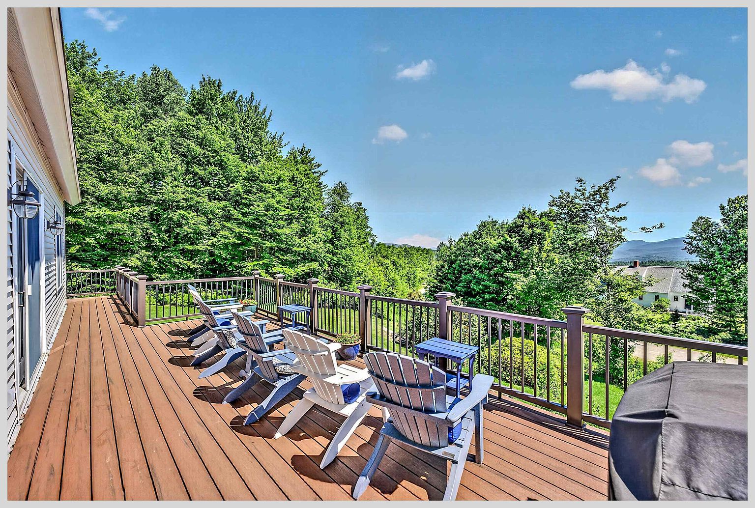 This spacious wooden deck offers a scenic vantage point overlooking a lush, tree-filled landscape and distant mountains. The area is furnished with several Adirondack chairs and small side tables, creating a comfortable space for outdoor relaxation. A dark railing encloses the deck, which connects directly to the home's interior through a sliding glass door.
