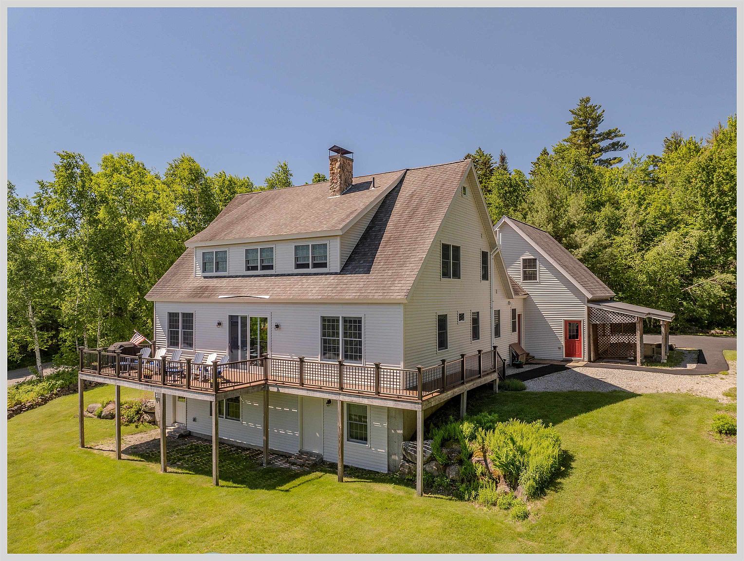 This elevated perspective captures the rear of a multi-level home featuring white horizontal siding and a prominent stone chimney. A spacious wooden deck extends from the main level, providing an ideal outdoor living space overlooking a lush, grassy lawn and surrounding mature trees. The property includes a secondary structure with a red door and a covered porch area, set against a clear blue sky.