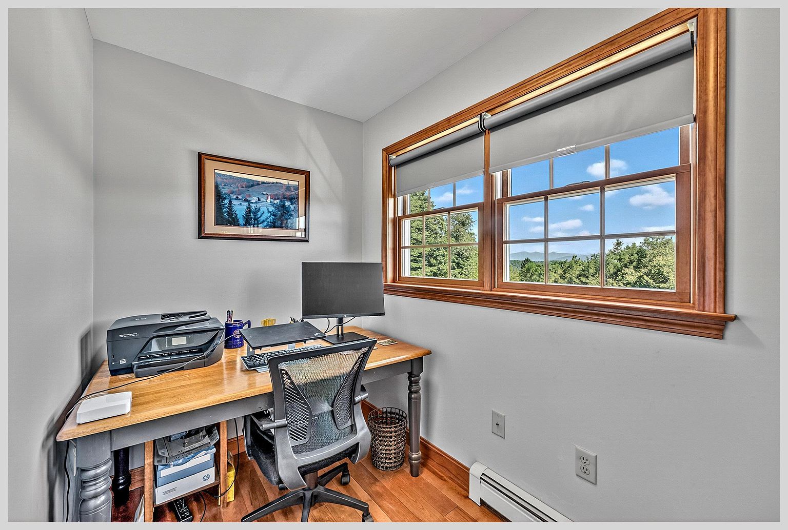 This compact home office features a wooden desk positioned against a light-colored wall, complete with a computer monitor, printer, and ergonomic chair. A large window with wooden trim provides a scenic view of the surrounding trees and distant mountains, filling the space with natural light. The room is finished with warm wood flooring and a framed landscape print, creating a quiet and productive workspace.