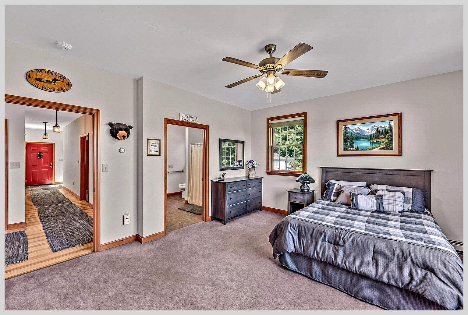 This bedroom features a cozy, rustic aesthetic with a plaid-patterned bedspread and a dark wood headboard. The room includes a matching dresser, a nightstand with a lamp, and a framed landscape painting above the bed, all set against neutral-toned walls and carpeted flooring. A doorway leads to an adjacent bathroom, while another opening provides a view into a hallway with a red front door.