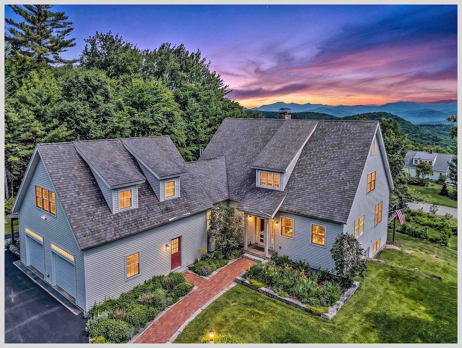 This elevated aerial perspective captures a charming, light-gray shingled home nestled against a lush, tree-lined backdrop with a stunning sunset view of distant mountains. The property features a multi-gabled roofline, a brick walkway leading to the front entrance, and an attached two-car garage. The well-manicured lawn and vibrant garden beds enhance the home's inviting curb appeal, creating a serene and picturesque setting.