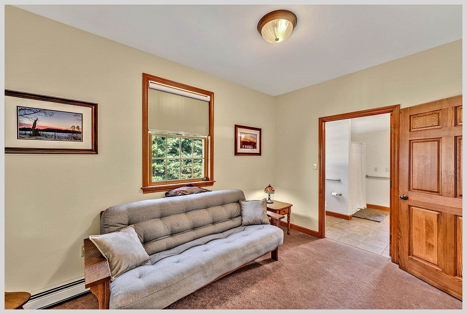 This cozy room features a neutral color palette with light-toned walls and brown carpeting, centered around a grey futon-style sofa. A window with a roller shade provides natural light, while a small side table with a decorative lamp adds a warm touch. The room includes a doorway leading into an adjacent bathroom, showcasing a functional layout suitable for a guest space or den.