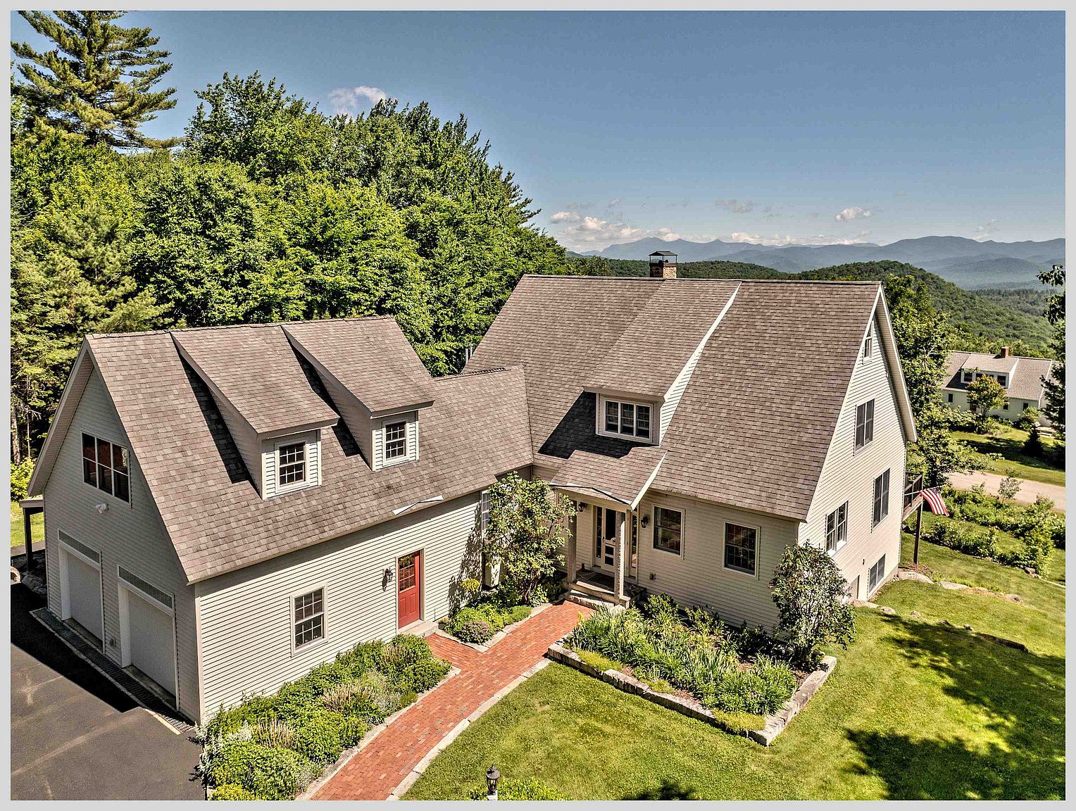 This elevated aerial perspective captures a charming, light-colored shingle-sided home nestled in a lush, mountainous landscape. The property features a multi-gabled roofline, a brick walkway leading to the front entrance, and an attached two-car garage, all surrounded by mature trees and manicured landscaping. The expansive mountain views in the background provide a serene and picturesque setting for this well-maintained residence.