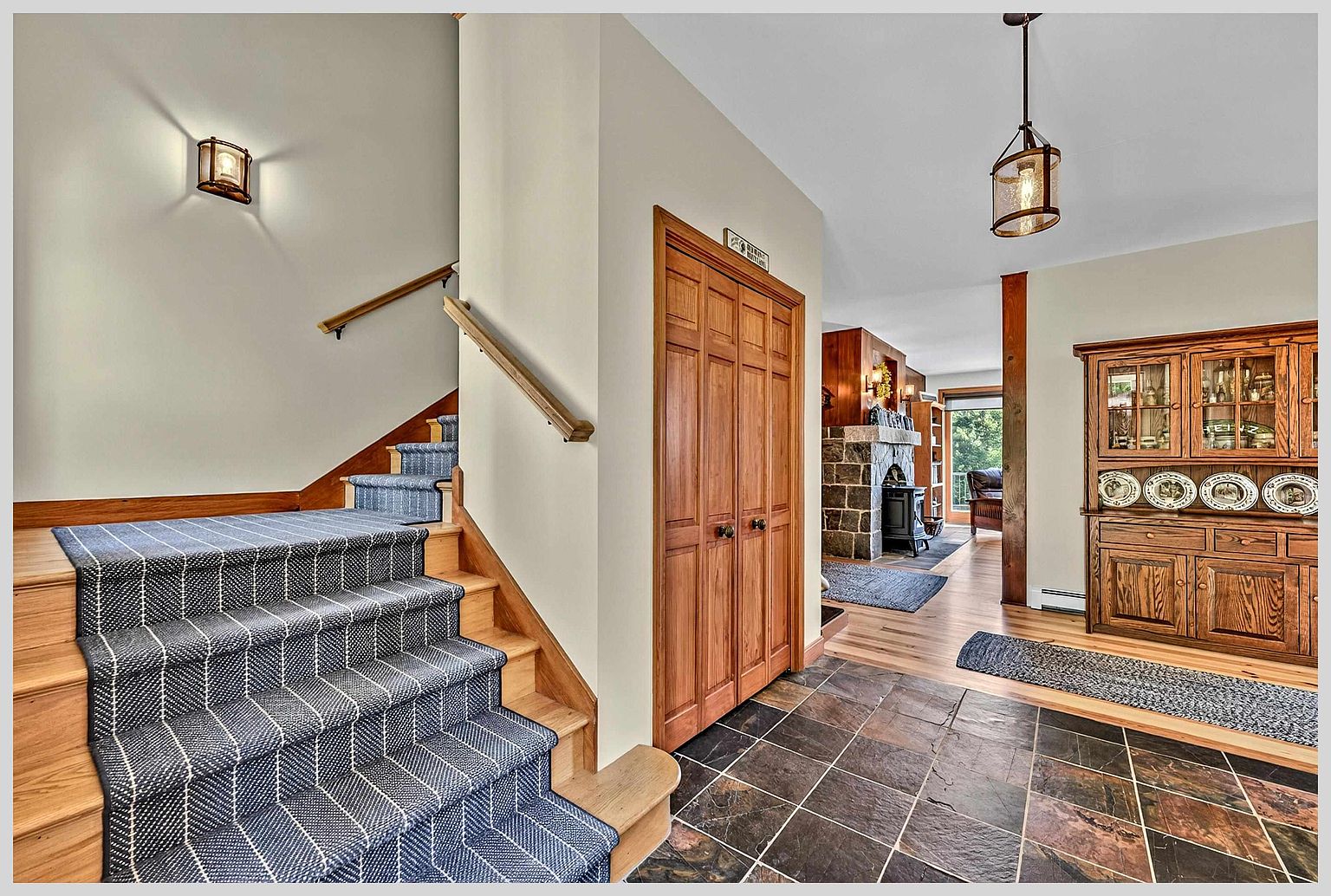This entryway features a staircase with a blue and white patterned runner leading to an upper level, adjacent to a wooden closet door. To the right, a slate-tiled floor transitions into a living area with a wooden hutch and a stone-clad chimney breast visible in the background. The space is illuminated by a hanging lantern fixture and a wall sconce, creating a warm and inviting atmosphere.