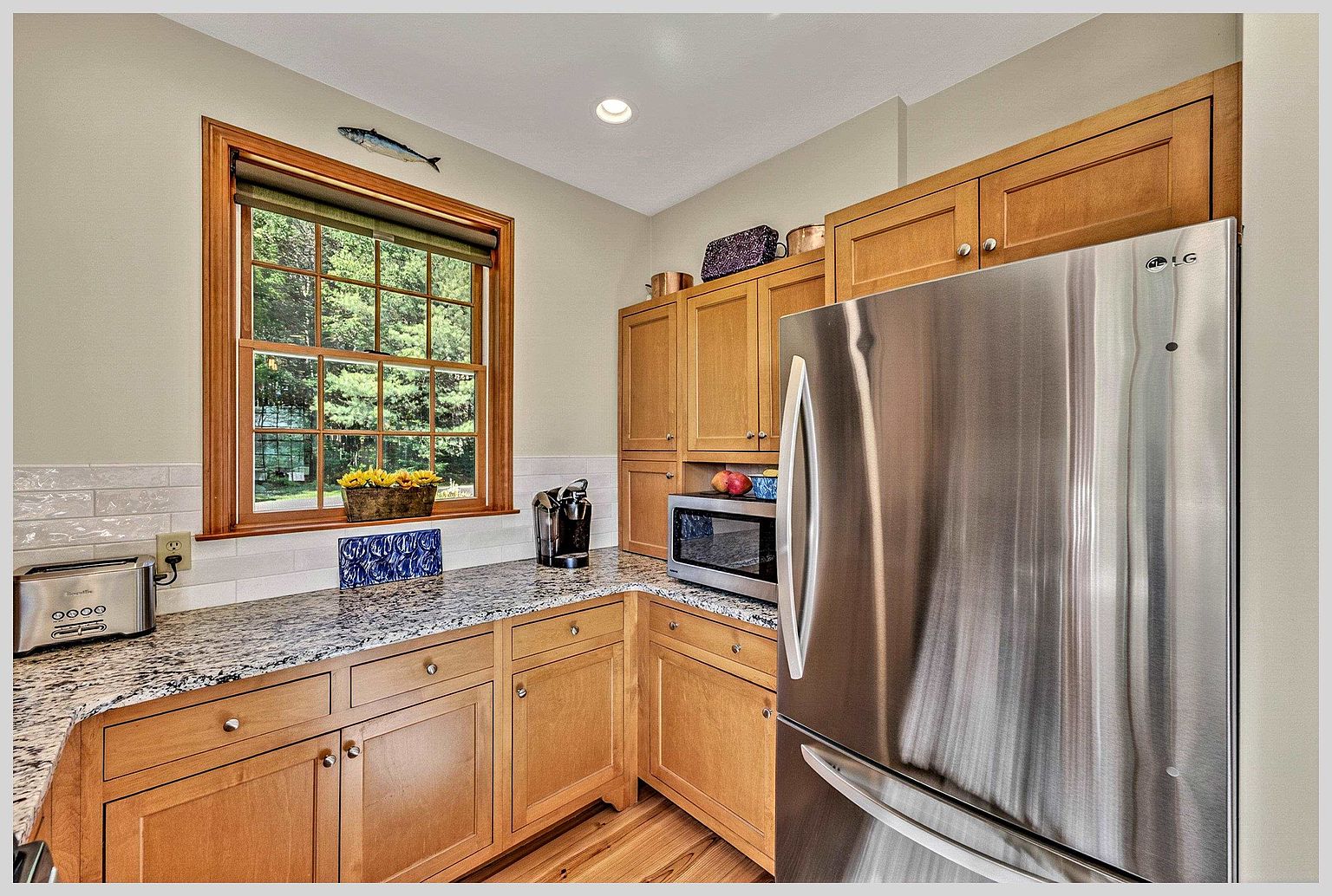 This kitchen features warm-toned wood cabinetry paired with speckled stone countertops and a white subway tile backsplash. A stainless steel refrigerator and microwave are integrated into the layout, while a window above the counter provides natural light and a view of the outdoors. The space is organized and functional, showcasing a classic, well-maintained aesthetic.