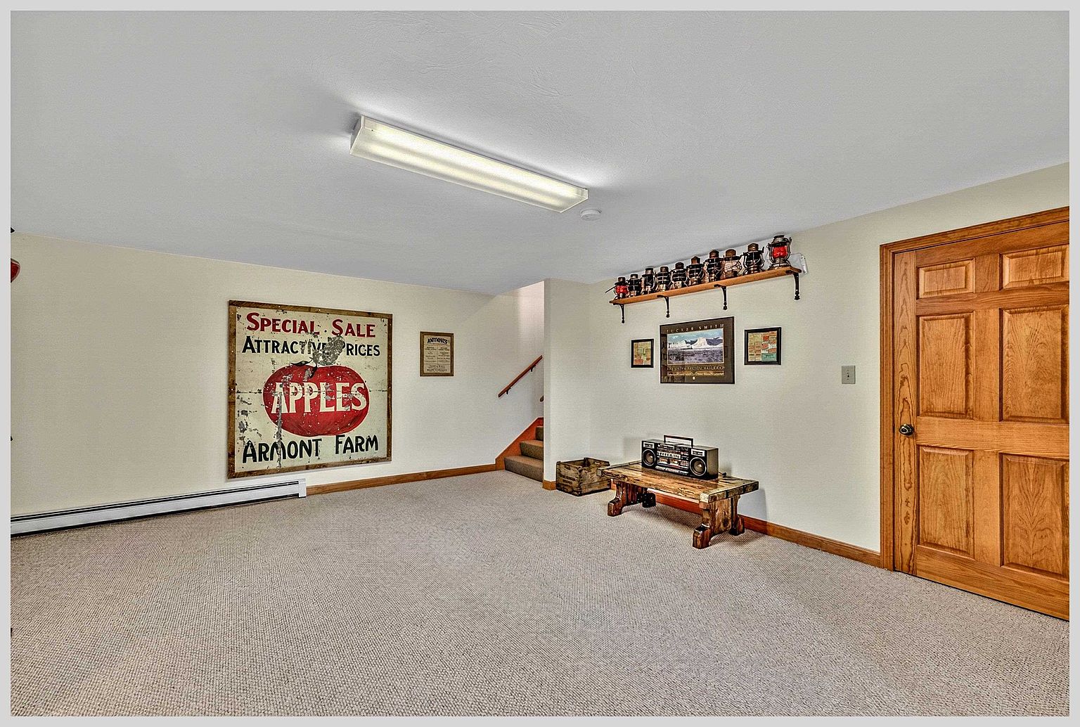 This finished basement space features neutral carpeting and light-colored walls, creating a versatile area for recreation or storage. The room is accented by a large vintage-style apple sign on the wall, a wooden bench holding a retro boombox, and a shelf displaying a collection of antique lanterns. A wooden door leads to another area, while a staircase in the background provides access to the upper levels of the home.