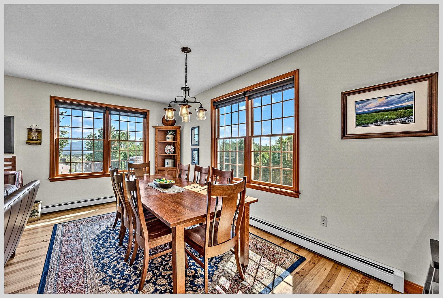 This bright and inviting dining room features a large wooden table surrounded by matching chairs, set atop a detailed patterned area rug. The space is illuminated by a multi-light chandelier and benefits from abundant natural light streaming through large windows with wood trim. A corner hutch and framed artwork on the walls add a touch of traditional charm to the warm, wood-floored room.