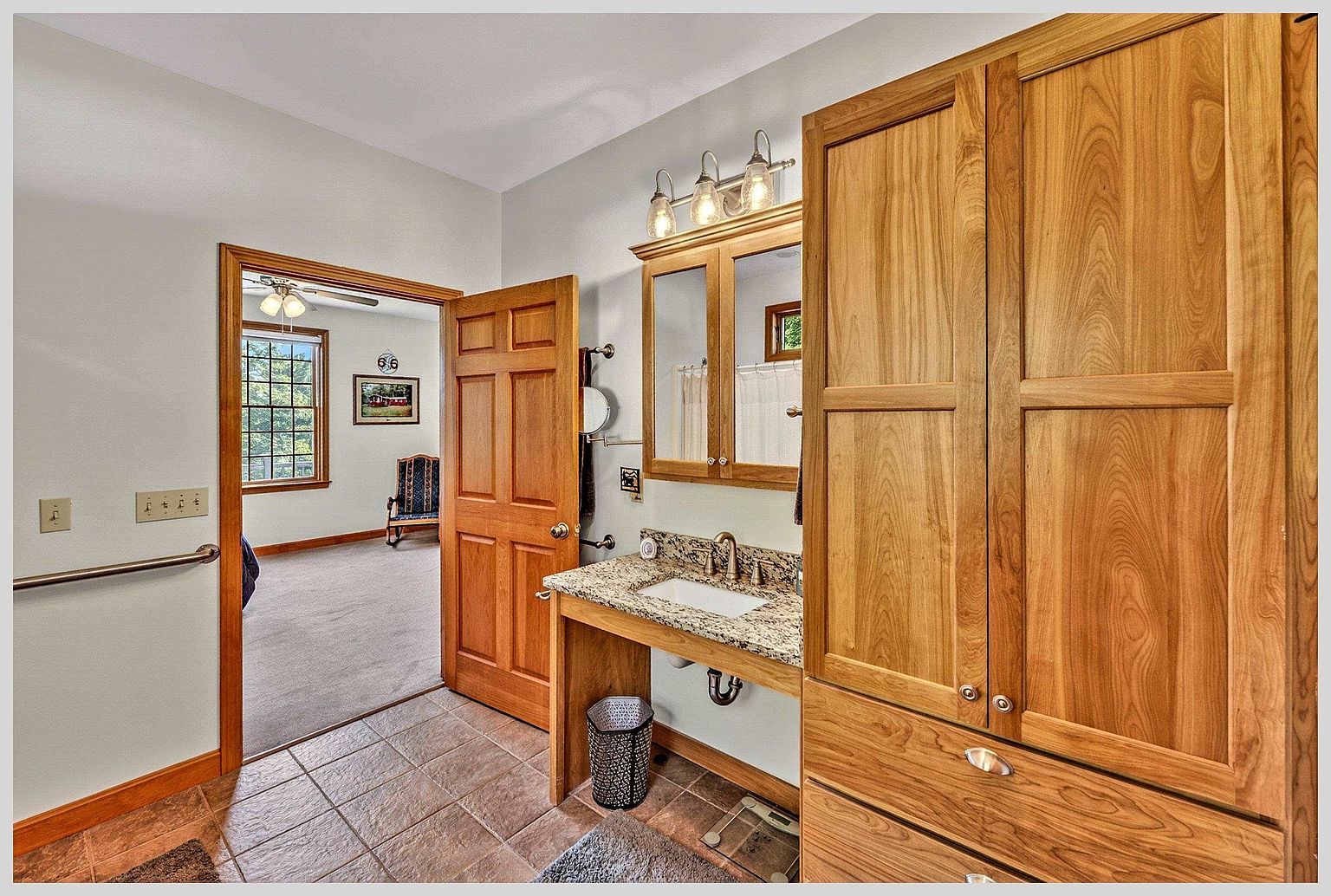 This bathroom features a vanity with a stone countertop and an open-concept base, paired with a tall, light-wood storage cabinet. A wooden door leads into an adjacent bedroom, which is visible through the doorway and contains a rocking chair and a window. The room is finished with neutral wall paint and tiled flooring, creating a functional and accessible layout.