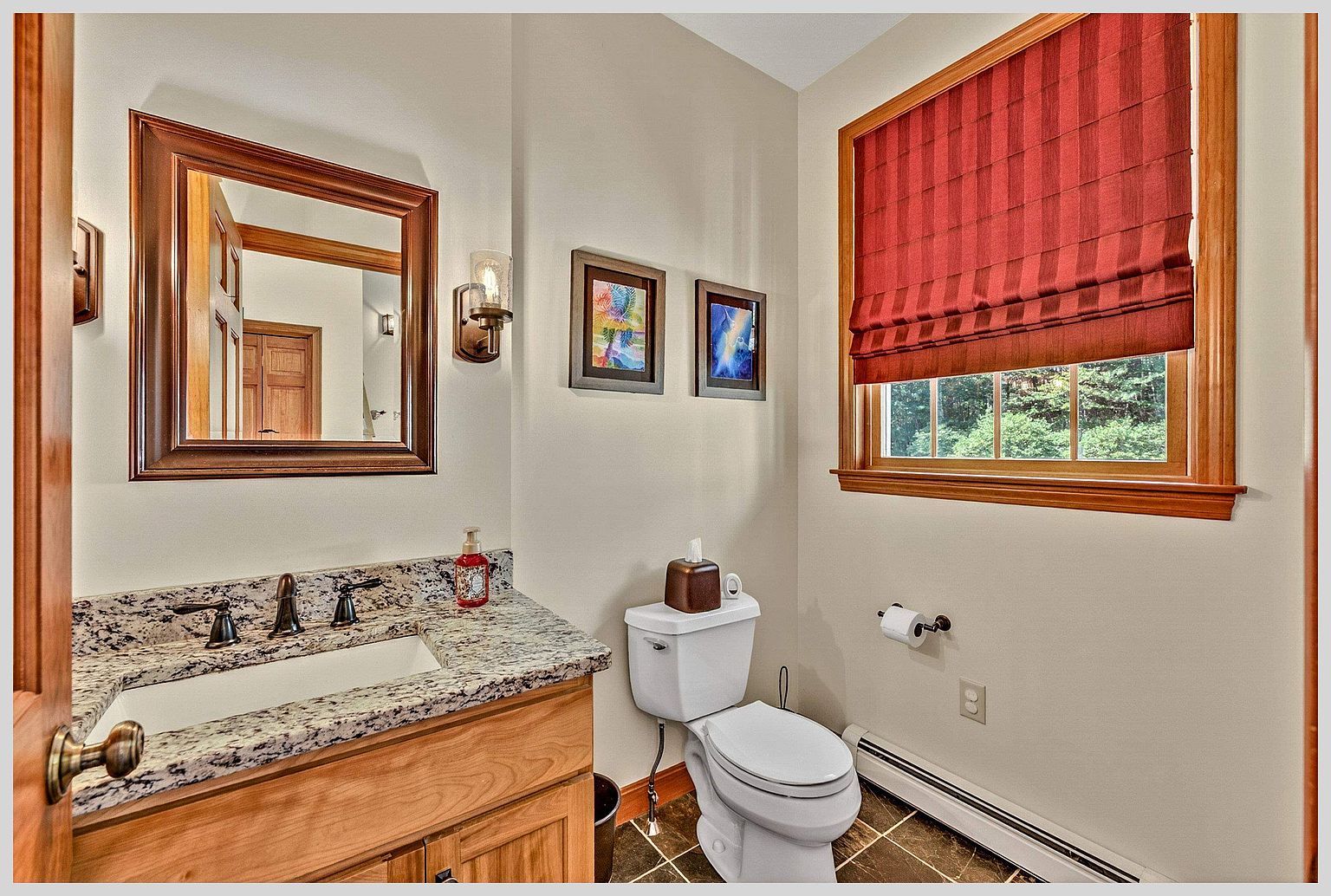This compact powder room features a vanity with a stone countertop and a framed mirror, complemented by warm-toned wood cabinetry. A white toilet sits beside a window dressed in a red fabric shade, while two framed artworks hang on the neutral-colored wall. The space is finished with dark, patterned floor tiles and a wall-mounted light fixture, creating a cozy and functional atmosphere.