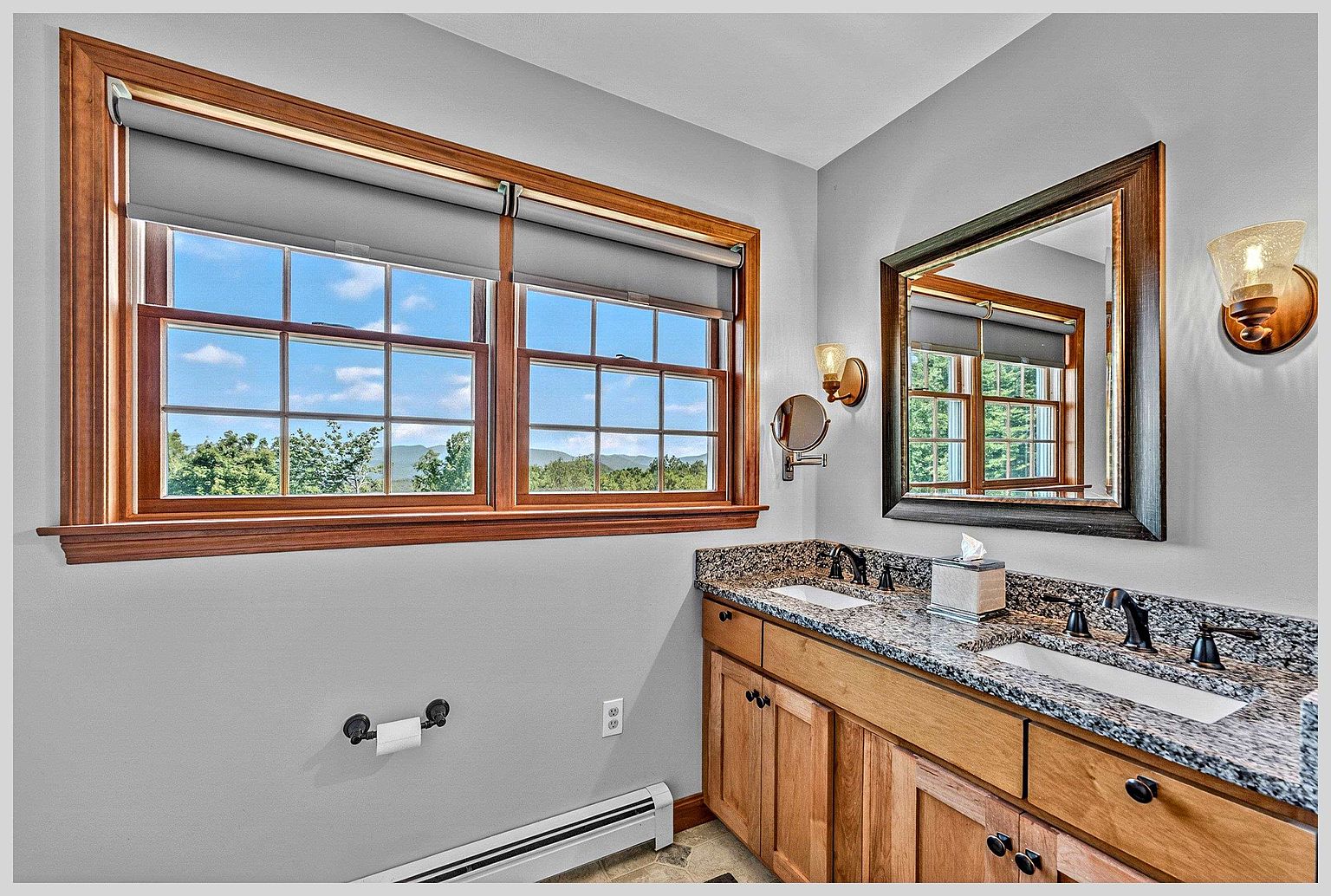 This bright bathroom features a dual-sink vanity with a speckled stone countertop and light wood cabinetry. A large, framed mirror hangs above the vanity, flanked by wall-mounted light fixtures, while a wide window with wooden trim provides natural light and a view of the outdoors. The space is finished with neutral gray walls and includes a wall-mounted toilet paper holder, creating a functional and well-lit atmosphere.