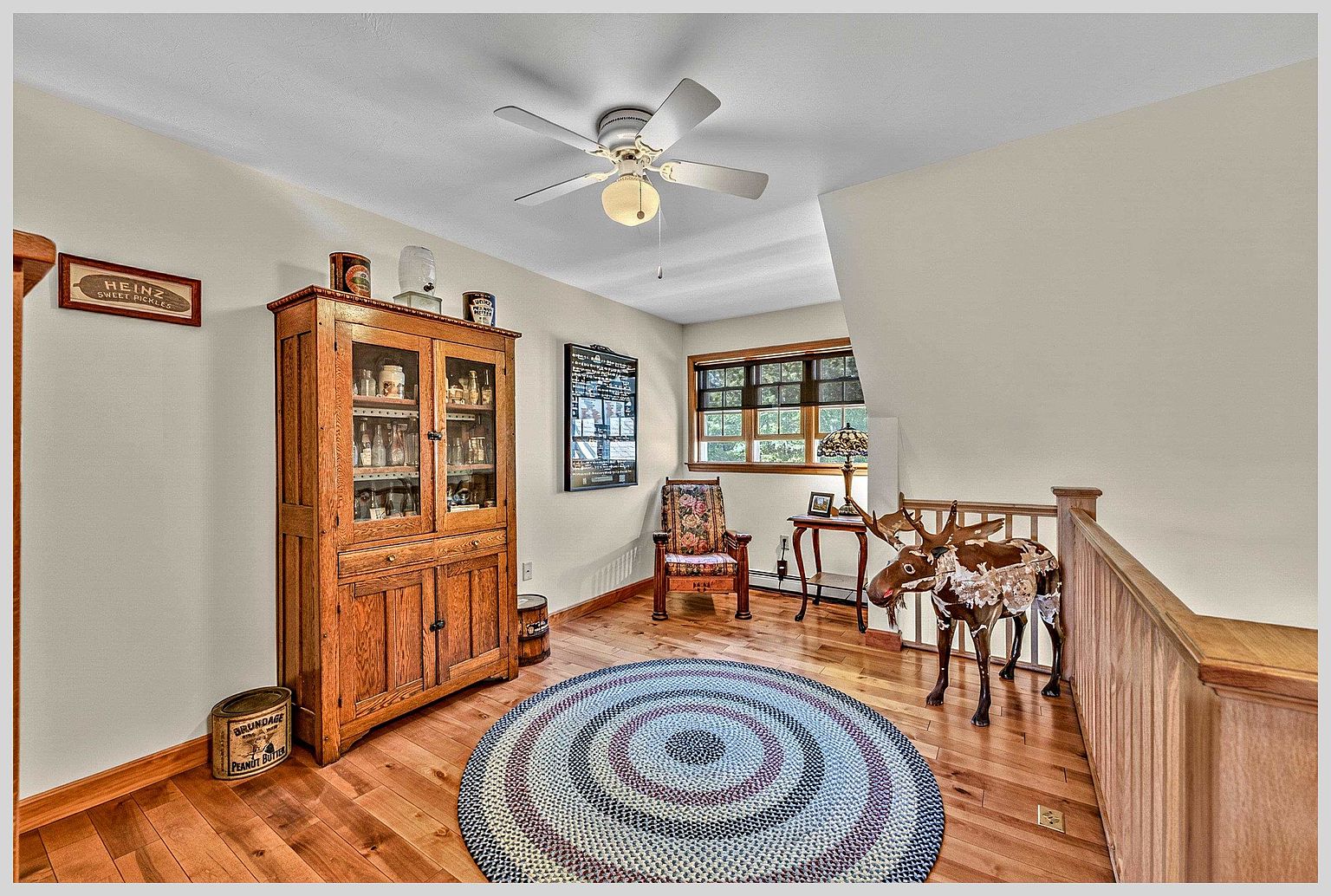 This cozy landing area features warm wood flooring and a circular braided rug, creating a welcoming and rustic atmosphere. The space is furnished with a tall wooden hutch, a floral-patterned armchair, and a decorative moose figure, all set against neutral walls. A ceiling fan with integrated lighting hangs above, while a window provides natural light to the area near the wooden staircase railing.