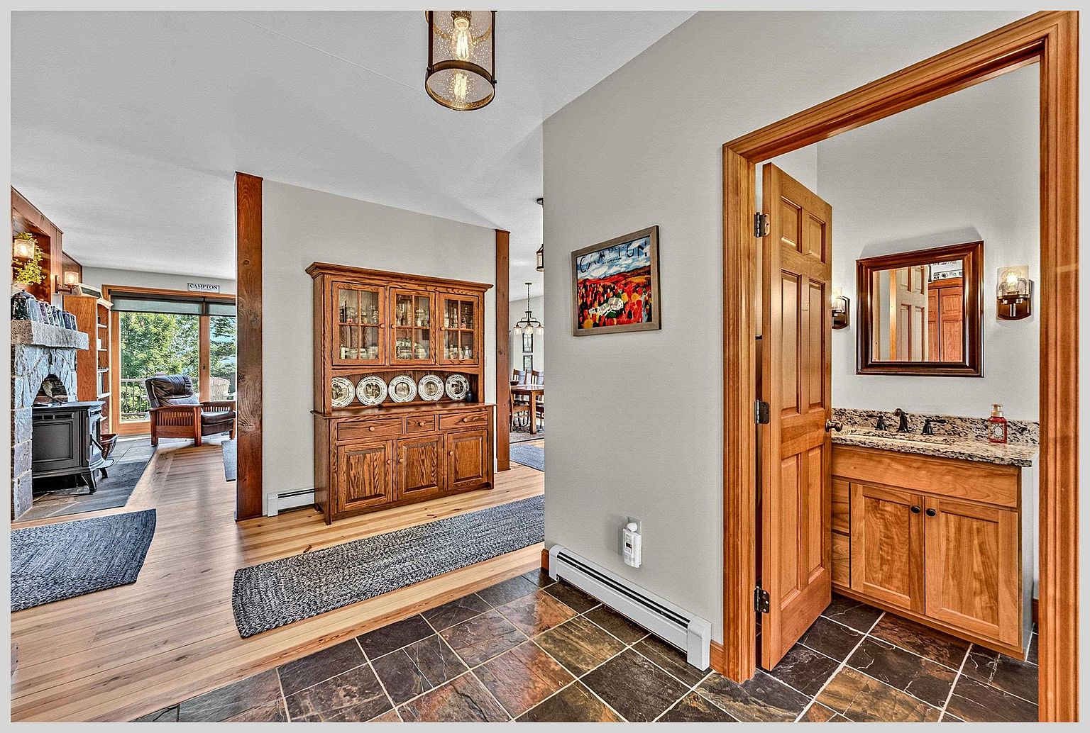 This interior view captures a transition space featuring a mix of slate-style tile flooring in the foreground and light wood flooring in the adjacent living area. A substantial wooden hutch stands against a neutral wall, while a doorway on the right reveals a bathroom with a stone-topped vanity and a framed mirror. The perspective looks through the home, highlighting the open floor plan, warm wood trim, and a rustic stone-clad chimney breast visible in the living room beyond.
