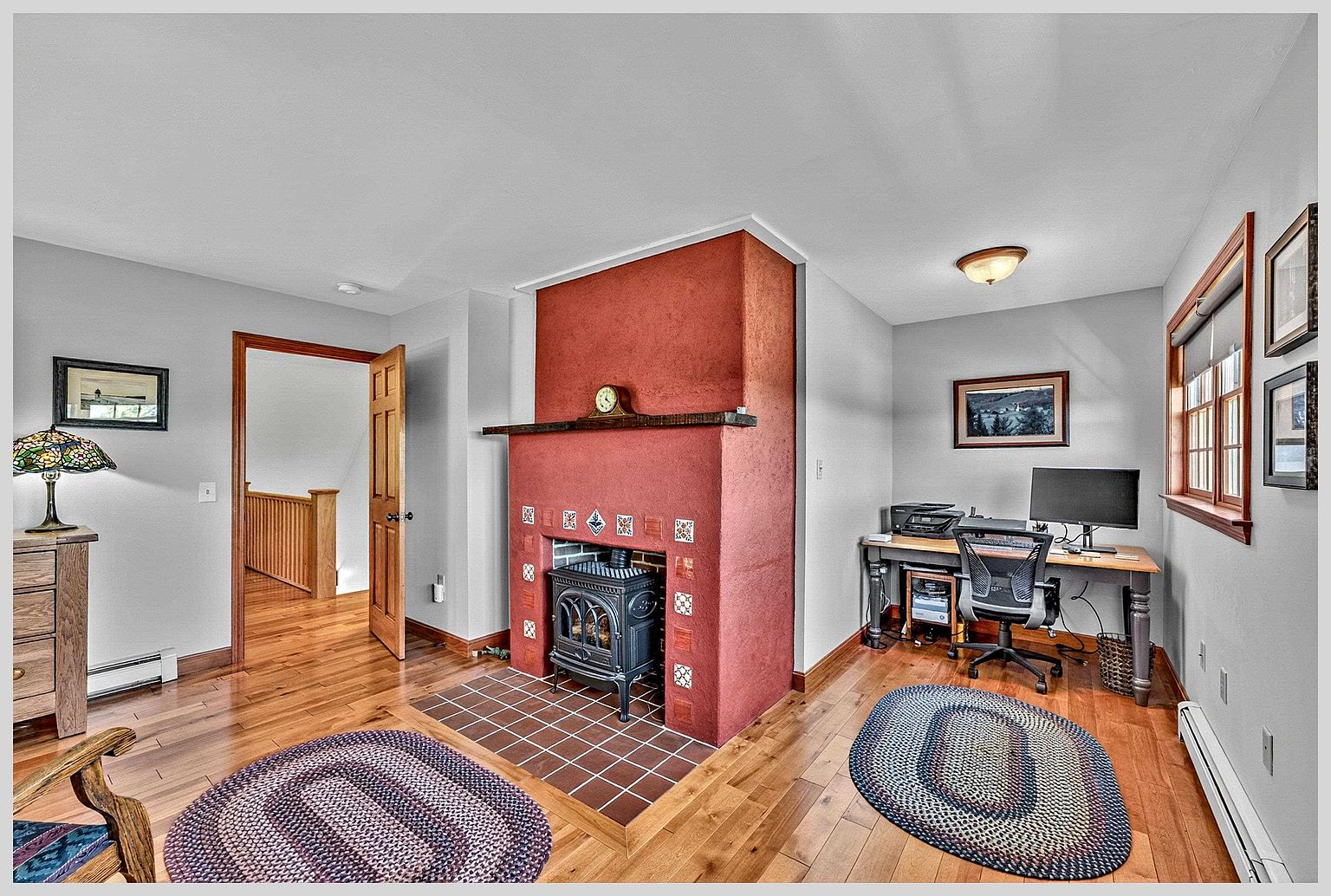 This versatile room features a prominent red-painted chimney breast with decorative tiles and a black wood-burning stove set on a tiled hearth. To the right, a dedicated workspace is arranged with a desk, computer, and office chair, while the left side opens into a hallway with hardwood floors. The space is accented by two round braided rugs and framed artwork, creating a cozy and functional multi-purpose living area.