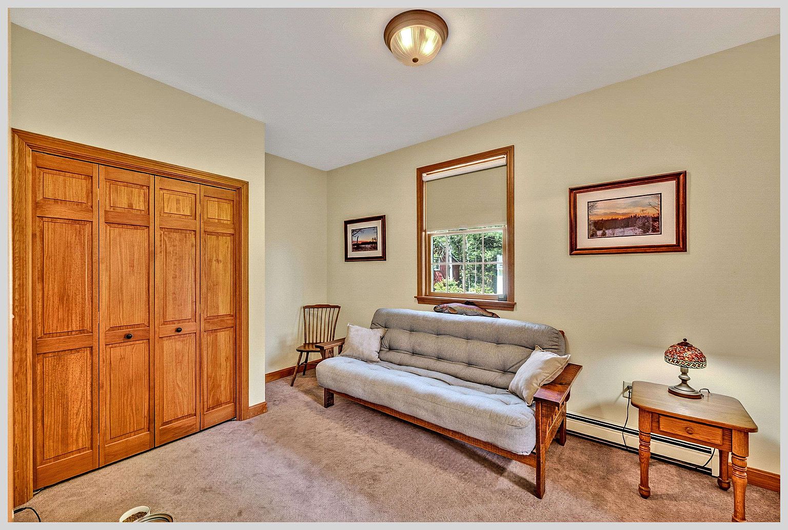 This cozy guest bedroom features neutral-toned walls, plush carpeting, and a wooden futon sofa that serves as a versatile seating or sleeping area. The room is accented by a wooden side table with a decorative lamp, a classic wooden chair, and two framed landscape prints on the walls. A large wooden closet with bi-fold doors provides ample storage, while a central window allows natural light to brighten the space.