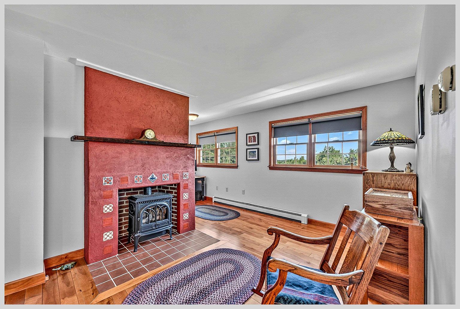 This cozy living room features a prominent red-textured chimney breast housing a black cast-iron wood stove set on a tiled hearth. The space is finished with warm-toned wood flooring, light-colored walls, and two large windows that provide ample natural light. A wooden rocking chair sits in the foreground, and the room is accented by braided oval rugs, creating a comfortable and inviting atmosphere.