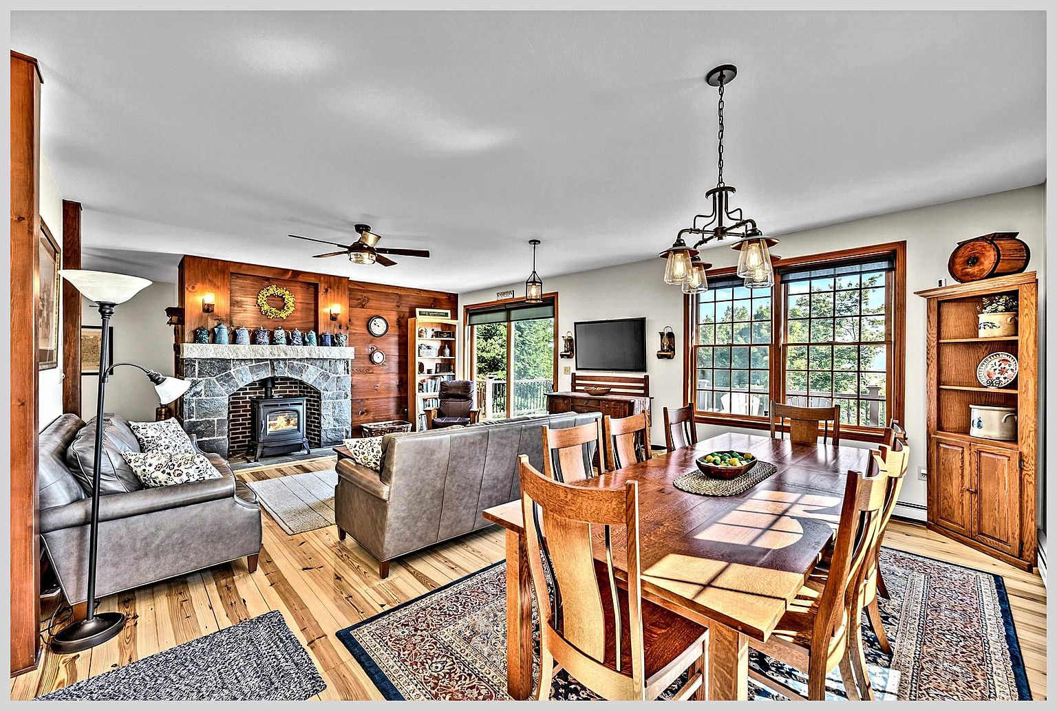 This open-concept living and dining area features warm wood-paneled walls and light-toned hardwood flooring throughout. A prominent stone-clad chimney breast with a wood-burning stove serves as the focal point of the living space, while a large wooden dining table anchors the adjacent dining area. The room is bathed in natural light from large windows and a sliding glass door, creating a cozy, rustic atmosphere.