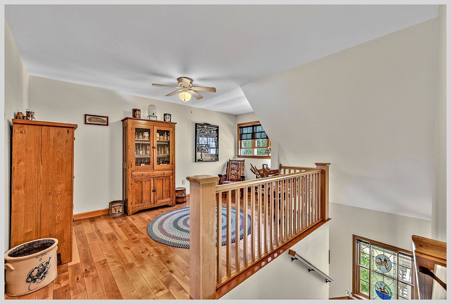 This view from the top of a staircase overlooks a landing area featuring warm-toned wood flooring and a classic wooden hutch. A round, multi-colored braided rug sits in the center of the space, while a wooden railing separates the landing from the stairwell. To the right, a stained-glass window adds a decorative touch to the stairwell, creating a cozy and traditional atmosphere.