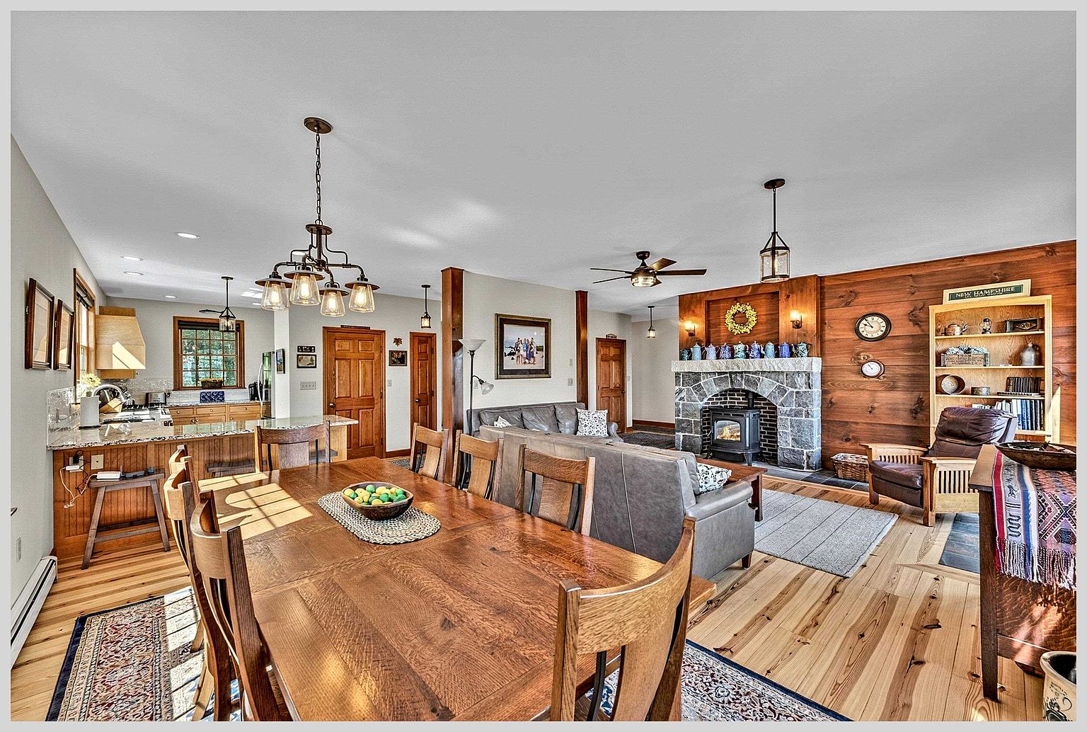 This open-concept living and dining area features warm wood flooring and a rustic aesthetic with wood-paneled walls. A stone-clad chimney breast serves as the focal point of the living space, while a large wooden dining table sits in the foreground. The room is illuminated by a combination of decorative pendant lighting and natural light, creating a cozy and inviting atmosphere.