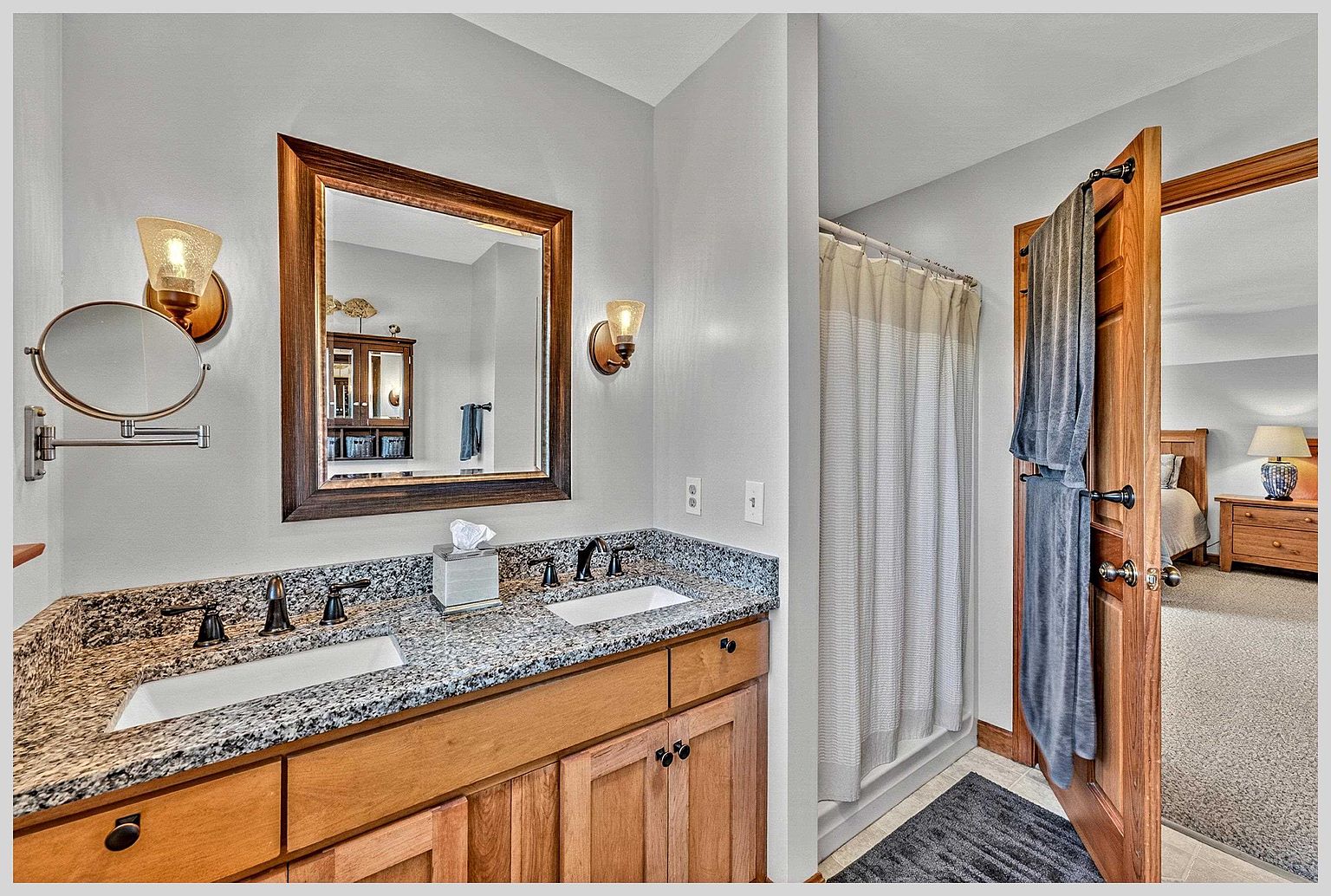This primary bathroom features a dual-sink vanity with a speckled stone countertop and light wood cabinetry. A large, framed mirror hangs above the vanity, flanked by two wall-mounted light fixtures, while a shower with a neutral curtain is positioned to the right. The open door reveals a glimpse into an adjacent bedroom, creating a sense of flow between the living spaces.