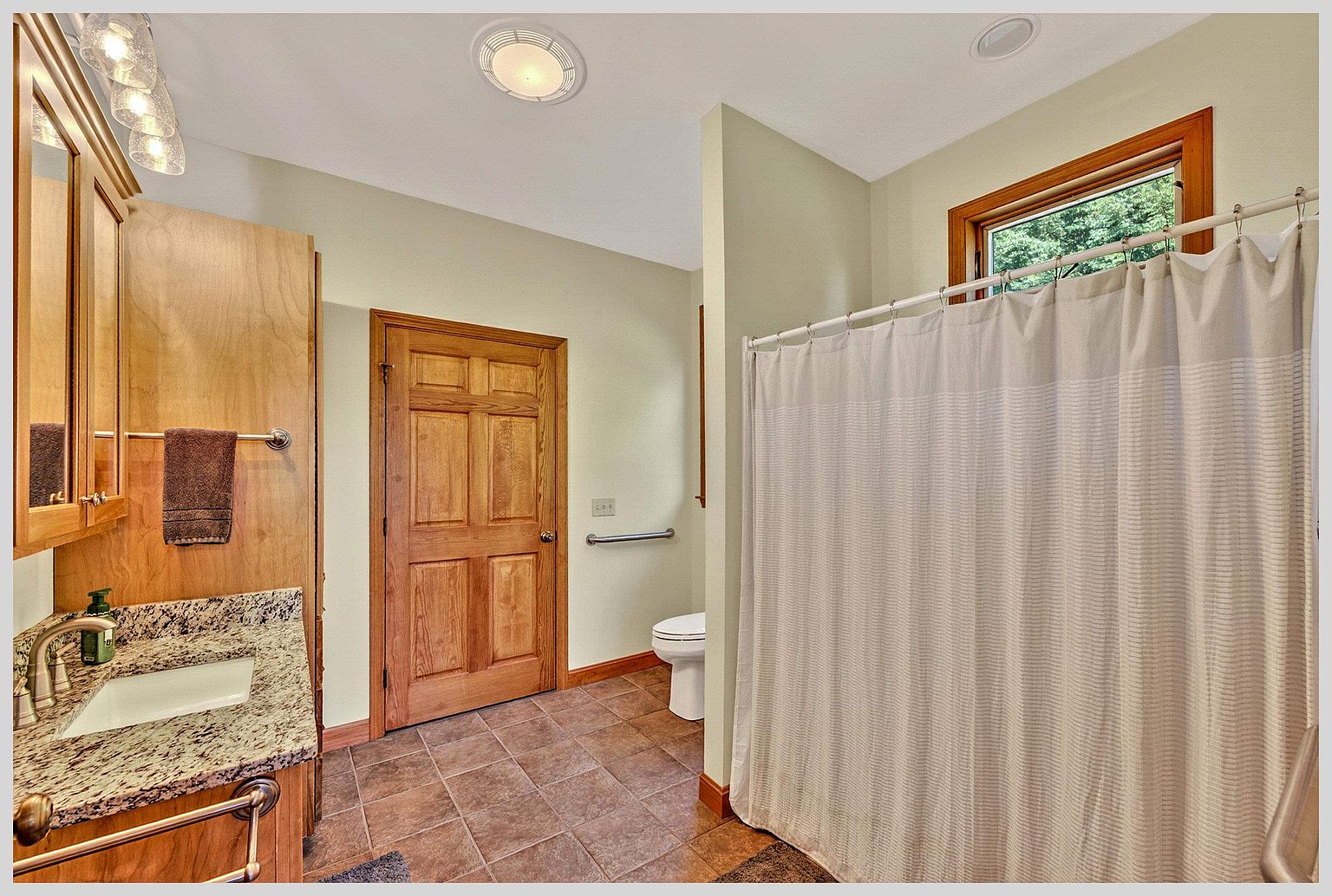 This bathroom features a vanity with a stone countertop, a light wood cabinet, and a matching mirror. A wooden door leads to a toilet area equipped with a grab bar, while a shower with a neutral-toned curtain is positioned to the right. The room is finished with light-colored walls and brown floor tiles, creating a functional and accessible space.