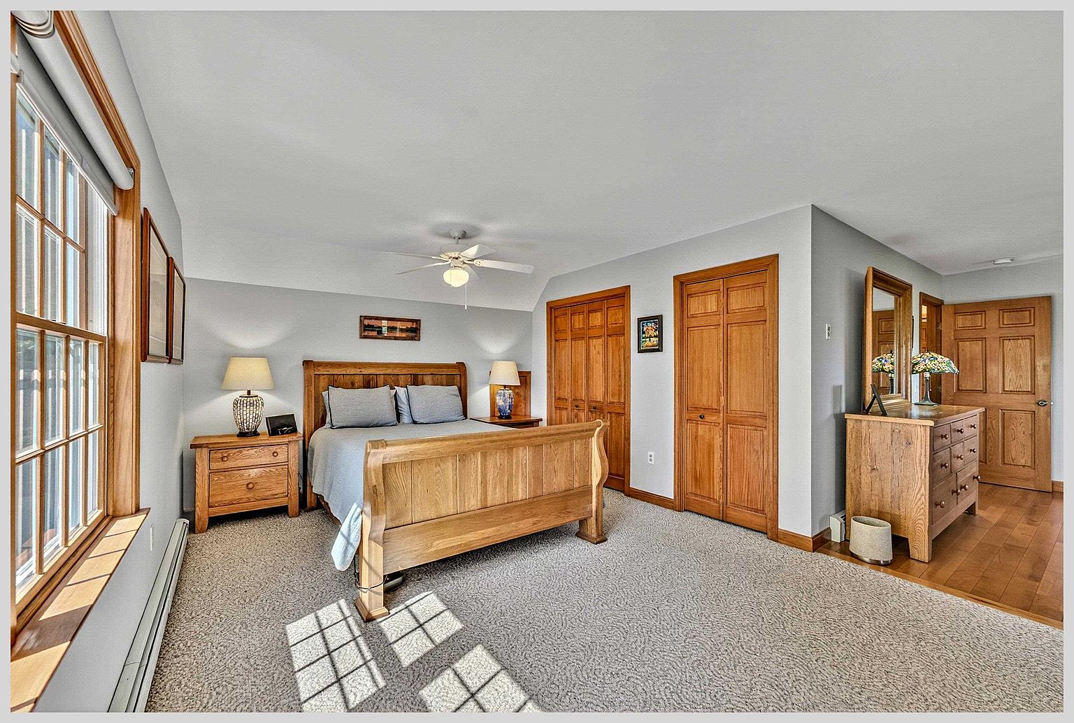 This spacious primary bedroom features a light wood bed frame and matching nightstands, complemented by a dresser with a mirror. The room is carpeted and includes two sets of wooden closet doors, with a large window on the left allowing natural light to fill the space. The neutral wall color and warm wood tones create a comfortable and inviting atmosphere.