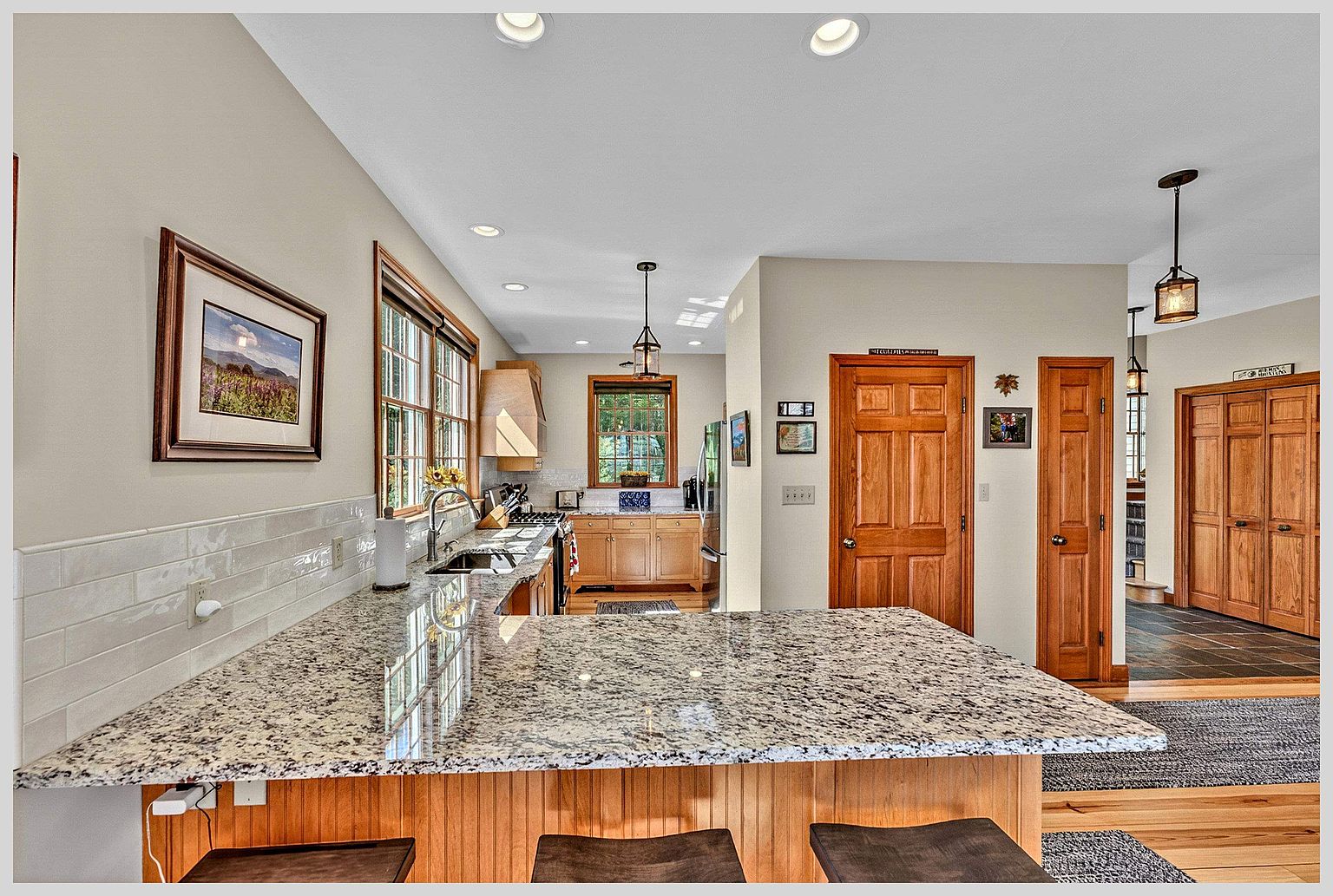 This kitchen features a spacious granite-topped island with bar seating, overlooking a functional workspace with light wood cabinetry and a tiled backsplash. The perspective captures the flow from the kitchen area into a hallway with warm wood doors and slate-style flooring. Recessed lighting and pendant fixtures provide a bright, inviting atmosphere throughout the open-concept space.