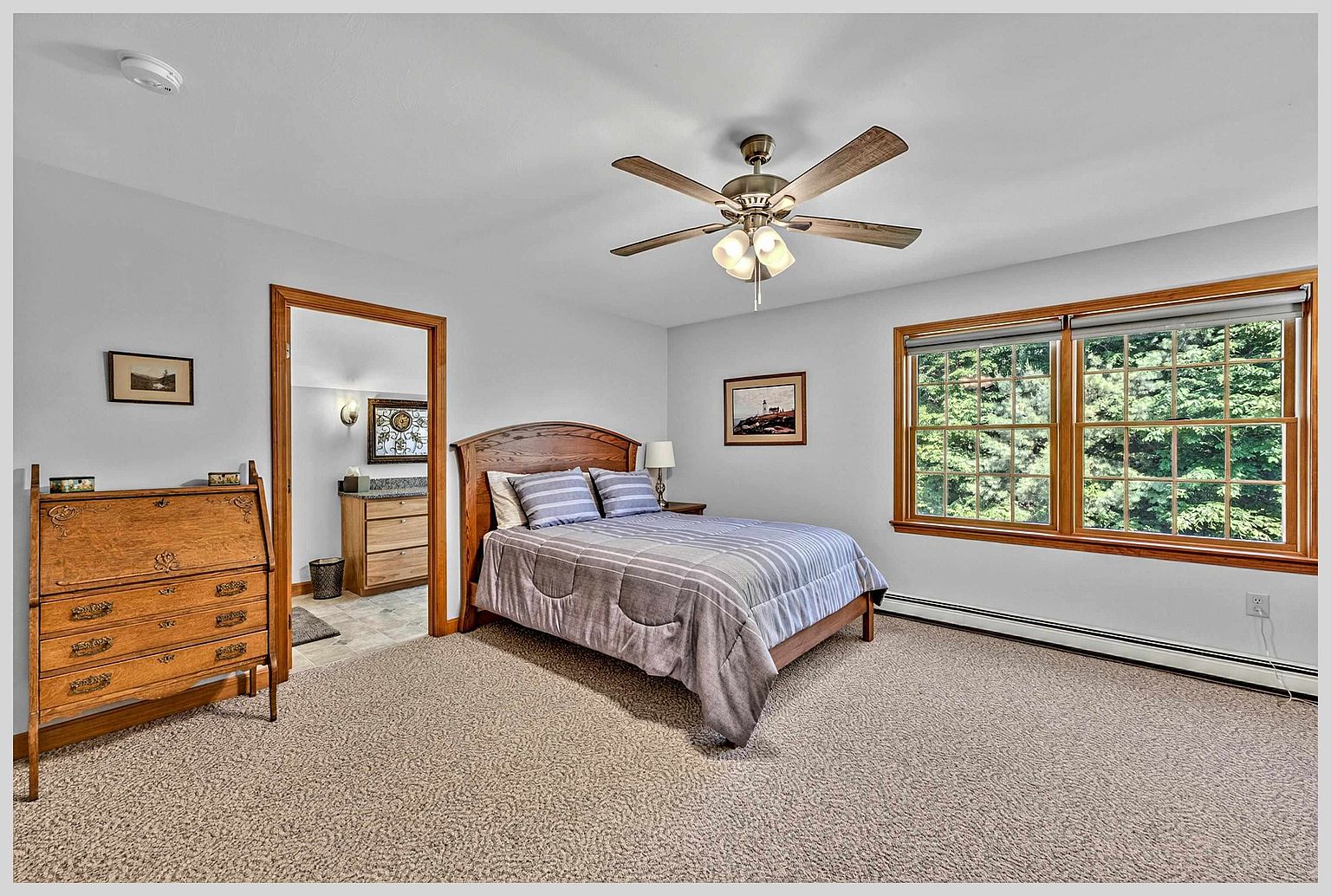 This bedroom features a wooden bed frame with a striped comforter and a matching wooden secretary desk positioned to the left. A doorway leads into an adjacent bathroom, while large windows on the right wall provide natural light and a view of the surrounding trees. The room is finished with neutral wall paint and wall-to-wall carpeting, creating a comfortable and traditional living space.