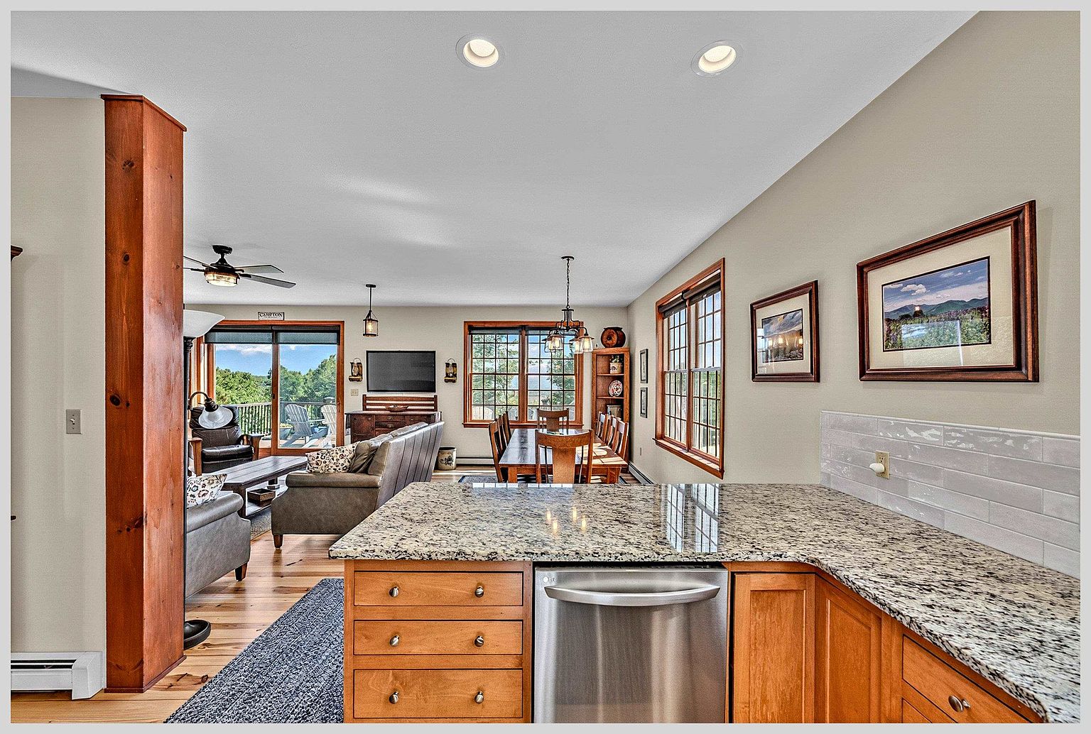 This open-concept living space features a kitchen in the foreground with a stone-topped island and stainless steel dishwasher, transitioning into a dining area and living room. The home showcases warm wood cabinetry, hardwood flooring, and a neutral color palette, with large windows and a sliding glass door that provide abundant natural light and views of the outdoors. The layout creates a cohesive, inviting atmosphere perfect for entertaining and daily living.