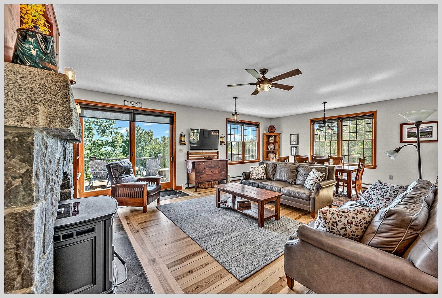 This open-concept living area features a warm, rustic aesthetic with light wood flooring and a prominent stone-clad chimney breast housing a wood stove. The space is furnished with comfortable leather seating, a wooden coffee table, and a dining area in the background, all bathed in natural light from large windows and a sliding glass door. The perspective captures the inviting flow between the living and dining zones, highlighting the cozy, mountain-lodge atmosphere.