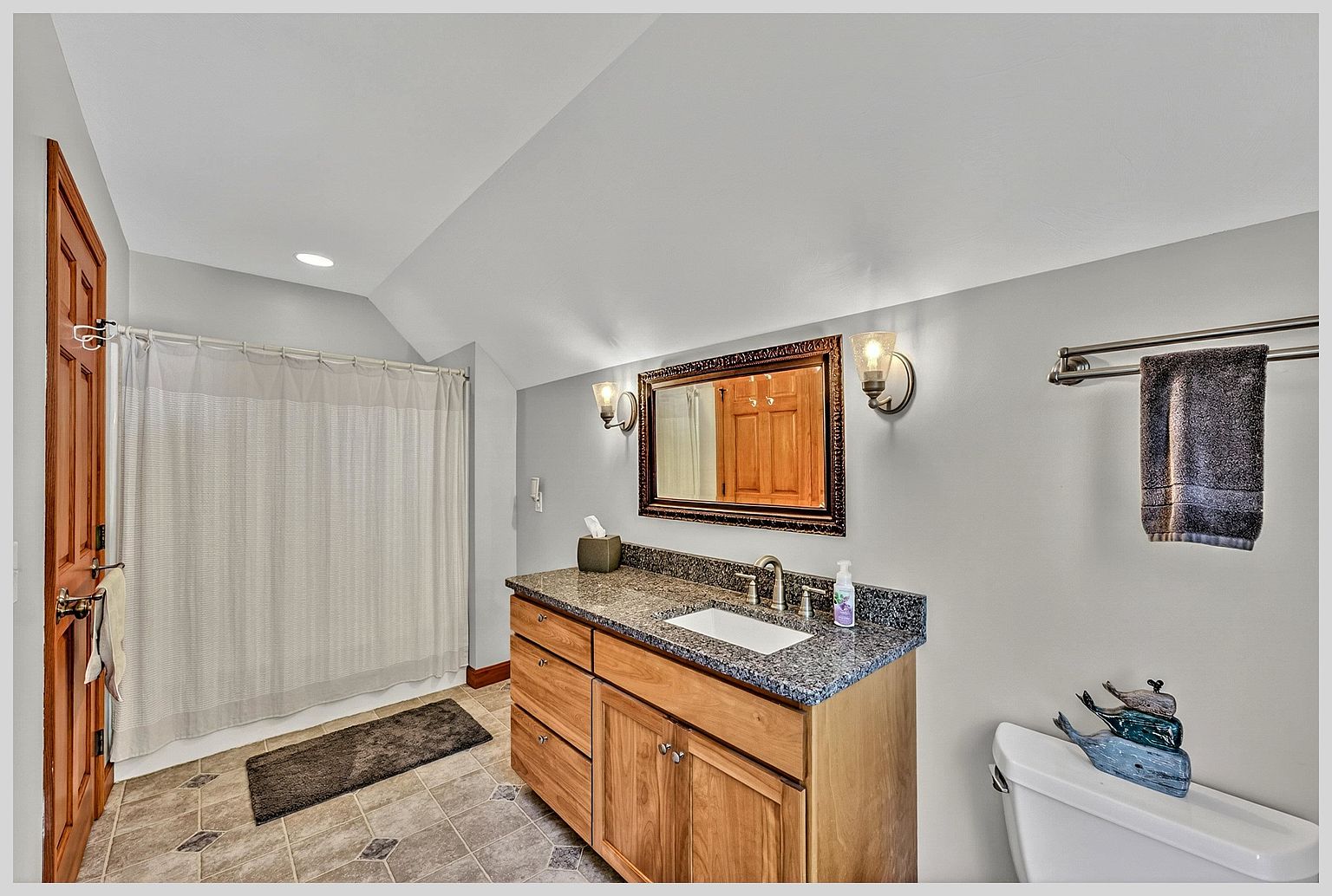 This bathroom features a light wood vanity topped with a dark, speckled stone countertop and a rectangular undermount sink. A framed mirror hangs between two wall-mounted light fixtures, while a shower with a light-colored curtain is positioned to the left. The room is finished with neutral gray walls and patterned tile flooring, creating a functional and clean aesthetic.