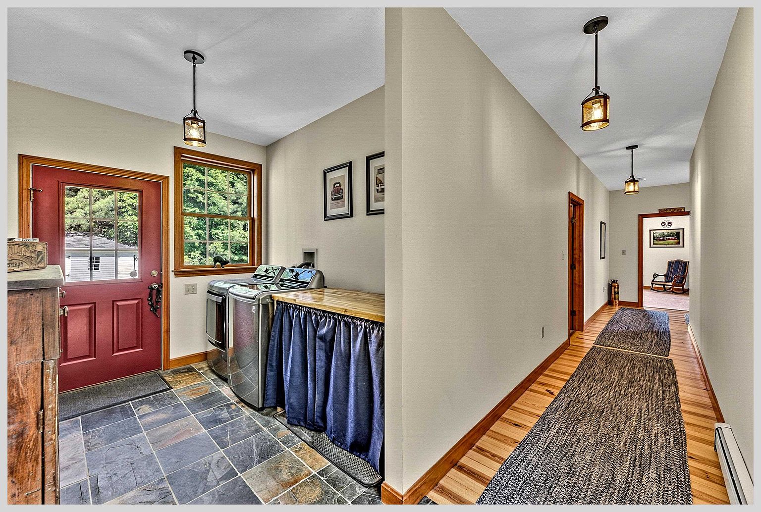 This functional space serves as a combined laundry room and entryway, featuring slate-style tile flooring and a vibrant red exterior door. A laundry station with a wooden countertop and a blue fabric skirt sits beside a window, while a transition leads into a bright hallway with light wood flooring and two pendant lights. The perspective captures the seamless flow between the utility area and the home's interior circulation path.