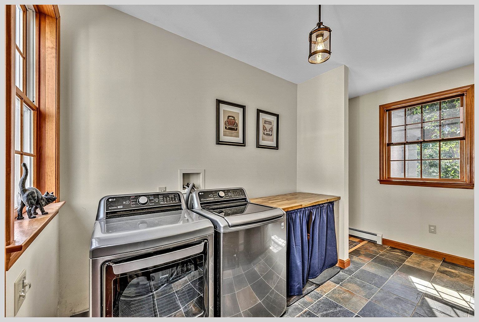 This functional laundry room features a pair of stainless steel LG appliances set against a neutral wall, complemented by a wooden countertop workspace with a blue curtain underneath for storage. The room is illuminated by a single pendant light fixture and includes slate-style floor tiles and wood-trimmed windows that bring in natural light. A decorative cat figurine sits on the windowsill, adding a touch of personality to this clean and organized utility space.