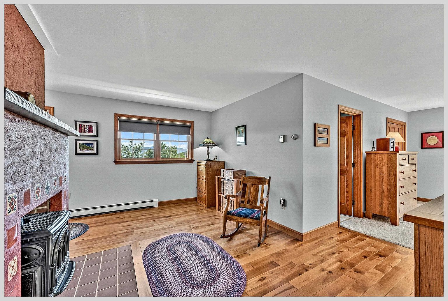 This cozy living area features light-toned wood flooring and a prominent wood-burning stove set against a textured, reddish-brown chimney breast. A wooden rocking chair sits in the center of the room near a braided oval rug, while a window provides natural light and a view of the outdoors. The space is accented by wooden furniture, including a chest of drawers and a small shelving unit, creating a warm and traditional atmosphere.