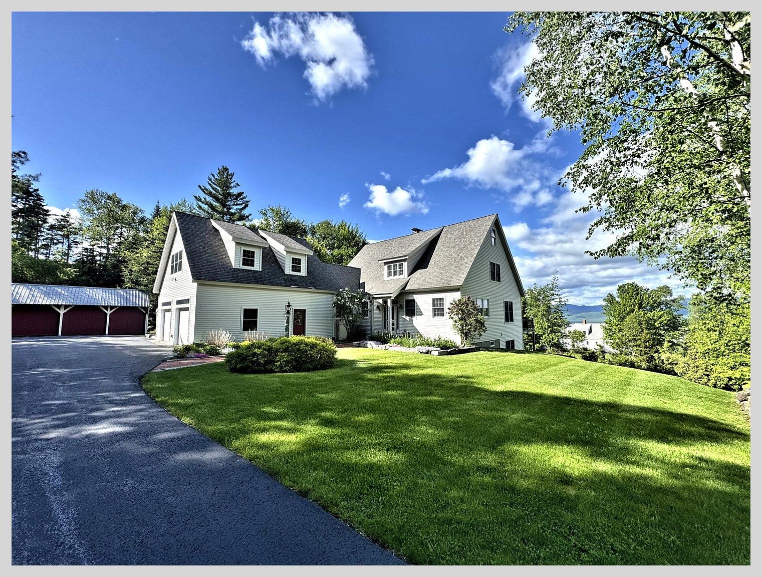 This charming, light-colored home features a classic gabled roofline with multiple dormer windows and a spacious, well-manicured front lawn. A long, paved driveway leads to an attached garage on the left, while mature trees frame the property against a bright blue sky with scattered clouds. The perspective captures the home from a low angle, emphasizing its inviting curb appeal and scenic surroundings.