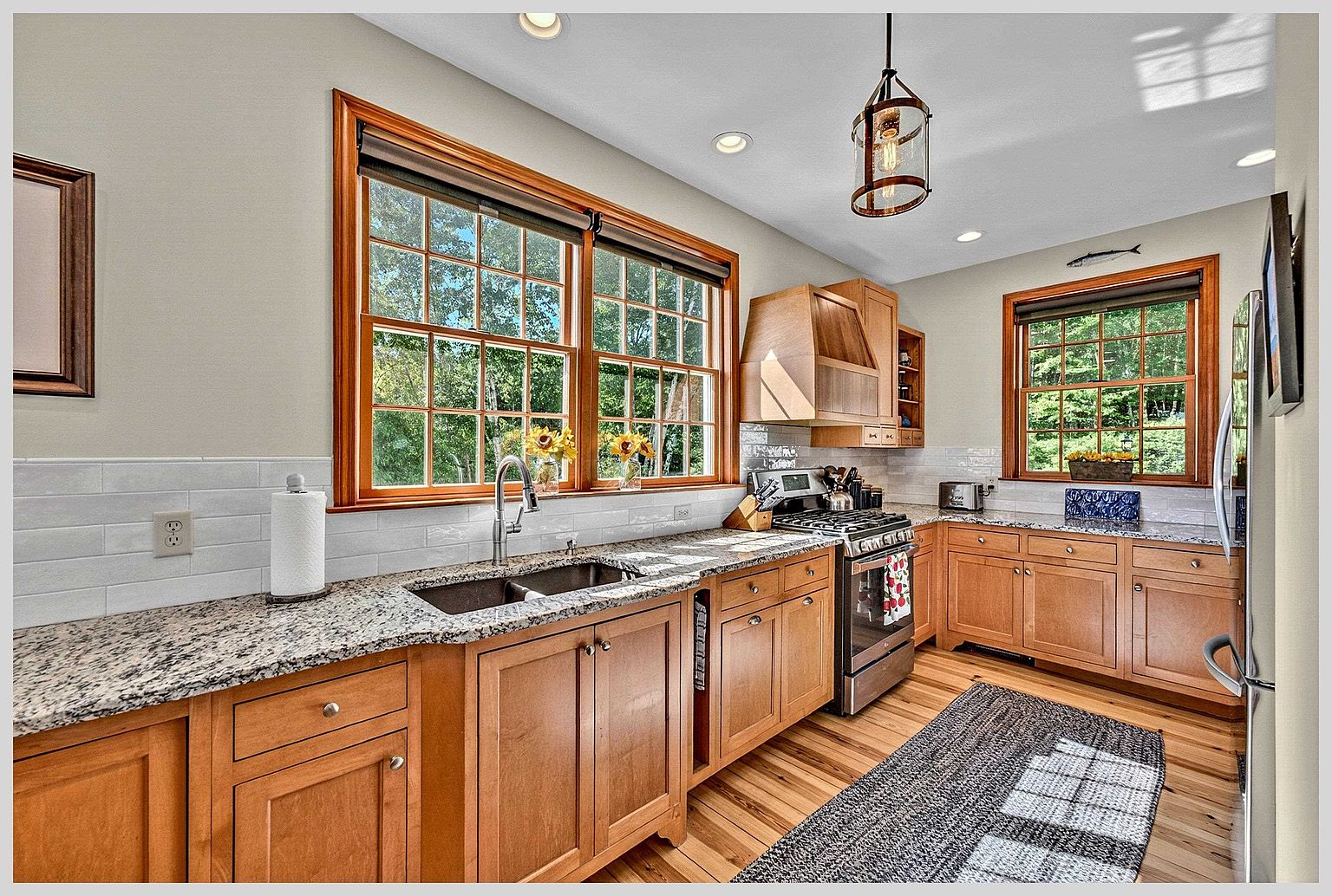 This bright and inviting kitchen features warm-toned wood cabinetry paired with speckled stone countertops and a white subway tile backsplash. The space is well-lit by a combination of recessed ceiling lights and a decorative pendant fixture hanging over the sink area, while large windows provide ample natural light and views of the outdoors. A stainless steel gas range and refrigerator complete the functional layout, which is anchored by a textured runner rug on the hardwood flooring.