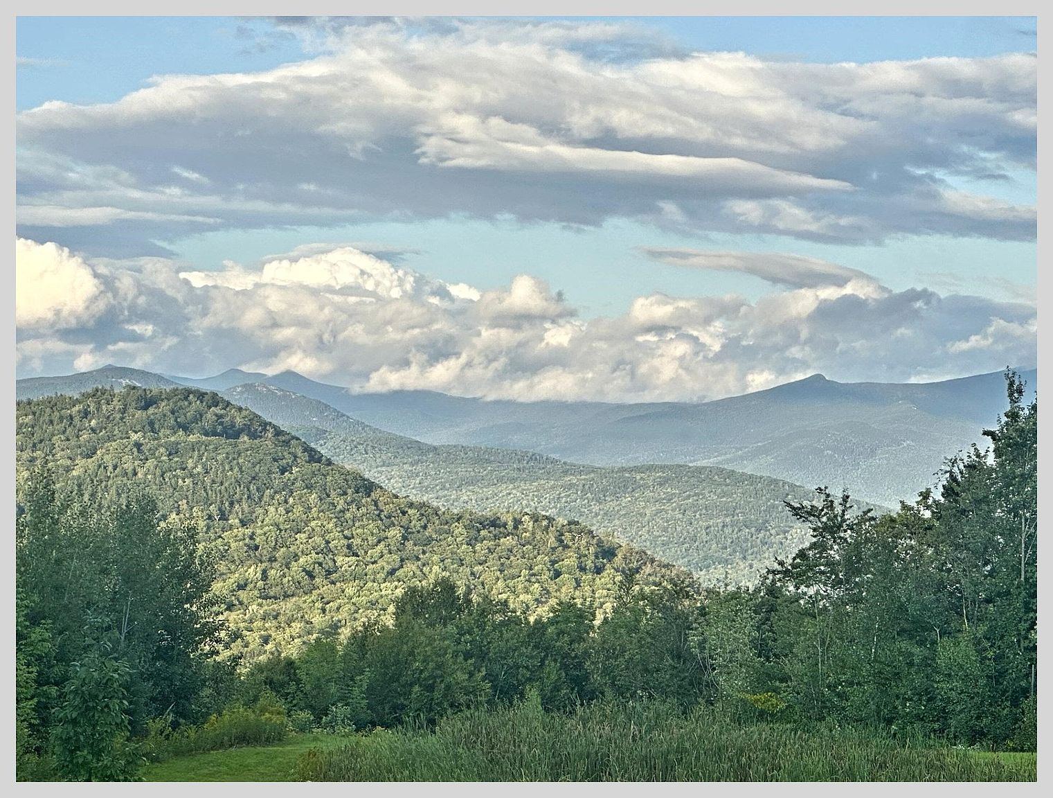 This image captures a scenic, expansive view of rolling mountain ranges under a sky filled with soft, layered clouds. In the foreground, a lush green meadow transitions into a dense forest of mature trees, creating a natural frame for the distant peaks. The perspective is elevated, offering a serene and picturesque outlook that highlights the property's natural surroundings and tranquil atmosphere.