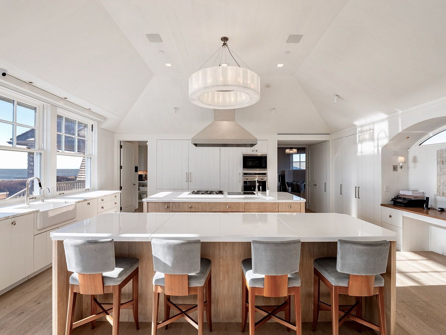 This bright and airy kitchen features a large central island with seating for four, complemented by a secondary island housing the cooktop and sink. The space is defined by clean white cabinetry, light wood accents, and a striking circular chandelier suspended from the vaulted ceiling. Large windows to the left provide a scenic view of the ocean, creating a serene and sophisticated atmosphere.