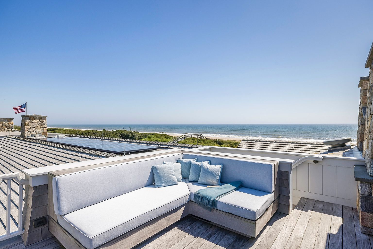 This elevated rooftop deck offers a serene, panoramic view of the ocean and coastline. The space is anchored by a custom L-shaped sectional sofa with light blue cushions and a matching throw, providing a comfortable vantage point for enjoying the coastal breeze. The deck features weathered wood decking, a stone-clad chimney breast, and a clear view of the adjacent roof with integrated solar panels.
