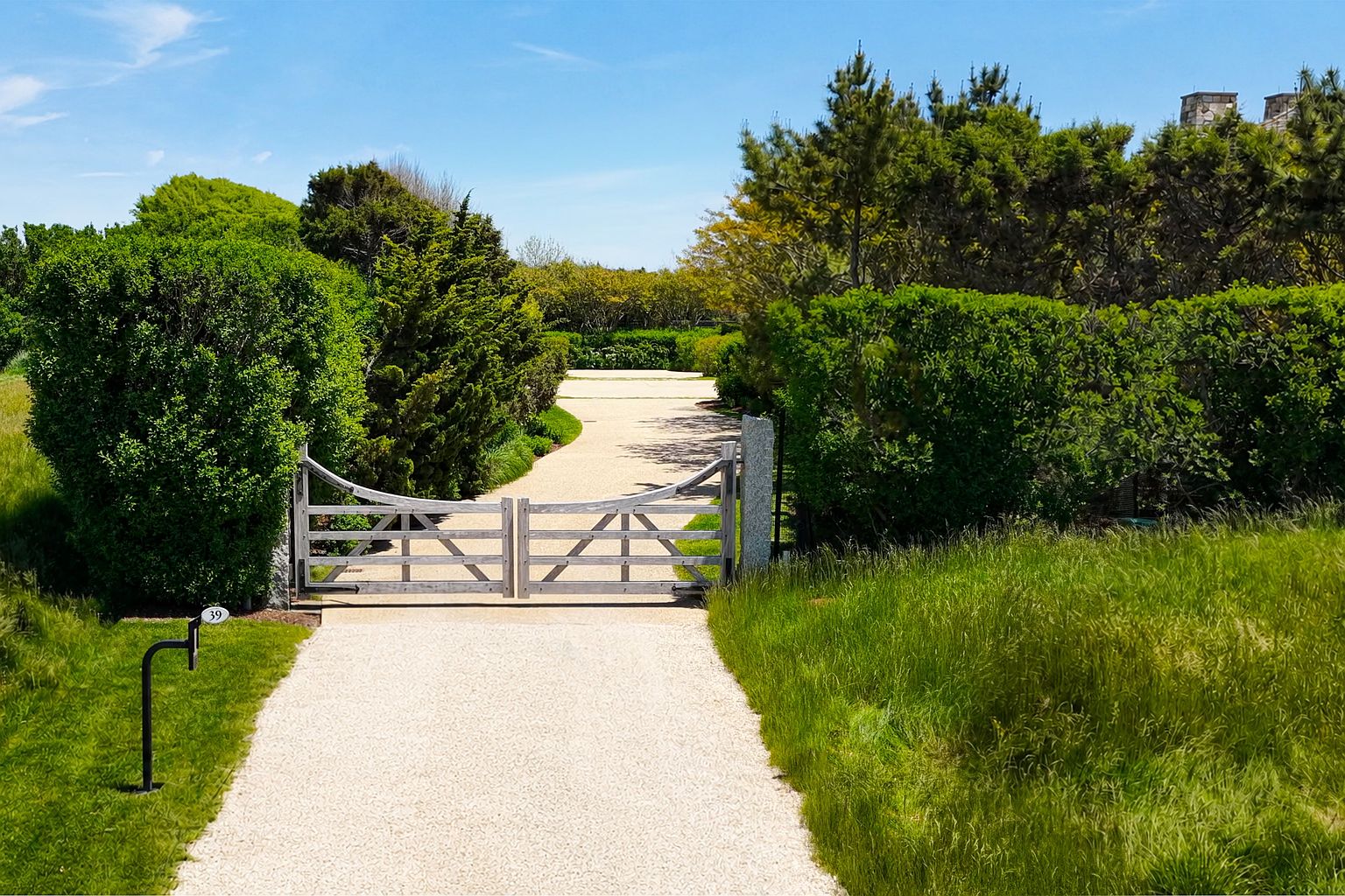 This image captures the gated entrance to a private property, featuring a light-colored gravel driveway that leads through a classic wooden gate. The entrance is framed by lush, mature hedges and greenery, creating a sense of privacy and seclusion. A small address sign marked '39' stands on a post to the left, and the perspective is a ground-level shot looking down the path toward the hidden residence.