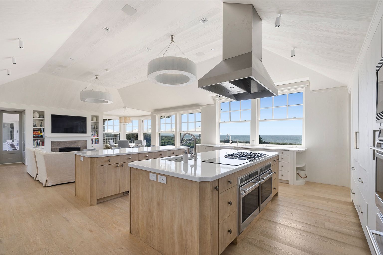 This expansive, open-concept kitchen features dual light wood islands with stone countertops and a prominent stainless steel range hood. The space is bathed in natural light from large windows that offer a panoramic view of the ocean, complemented by a vaulted ceiling with exposed beams. A cozy living area with a fireplace is seamlessly integrated into the same room, creating a bright and airy atmosphere perfect for entertaining.