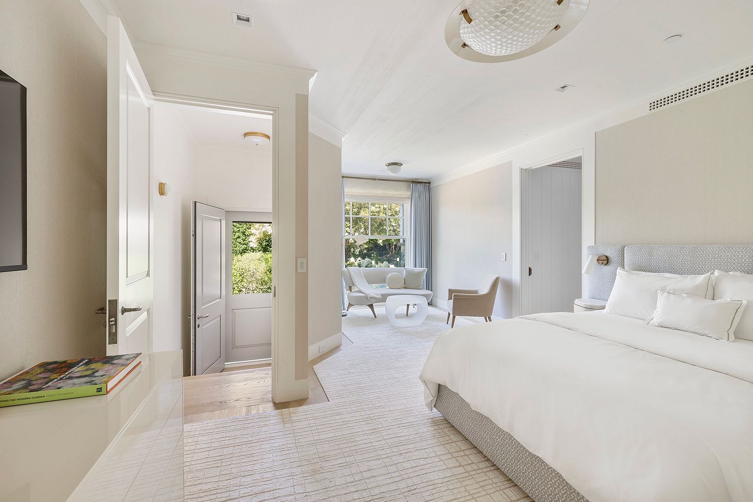 This bright and airy primary bedroom features a neutral color palette with a plush, textured bed and a cozy seating area near the window. The room flows seamlessly into a small entryway with a door leading to an outdoor space, creating a sense of openness and connection to the garden. The space is defined by soft carpeting, elegant lighting fixtures, and a clean, sophisticated aesthetic.