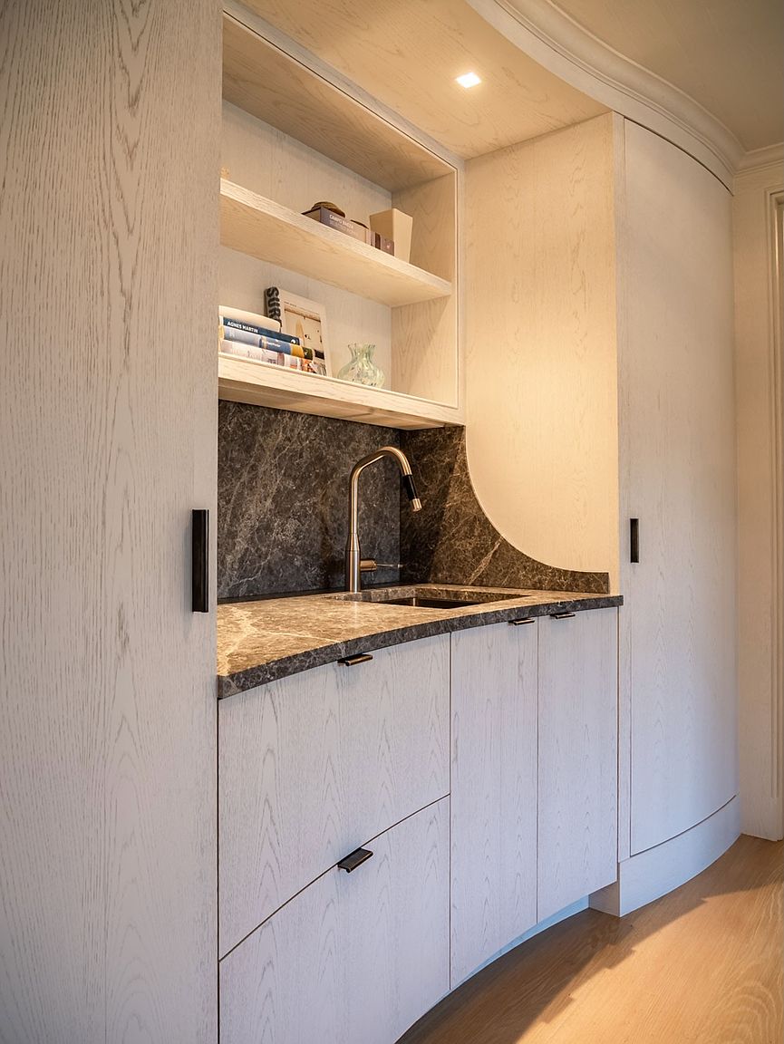 This elegant wet bar or kitchenette area features light wood cabinetry with a clean, minimalist aesthetic and matte black hardware. A dark, veined stone countertop and matching backsplash provide a sophisticated contrast, complemented by an integrated sink and faucet. The space includes open shelving for display and is illuminated by recessed lighting, creating a refined and functional transition area.