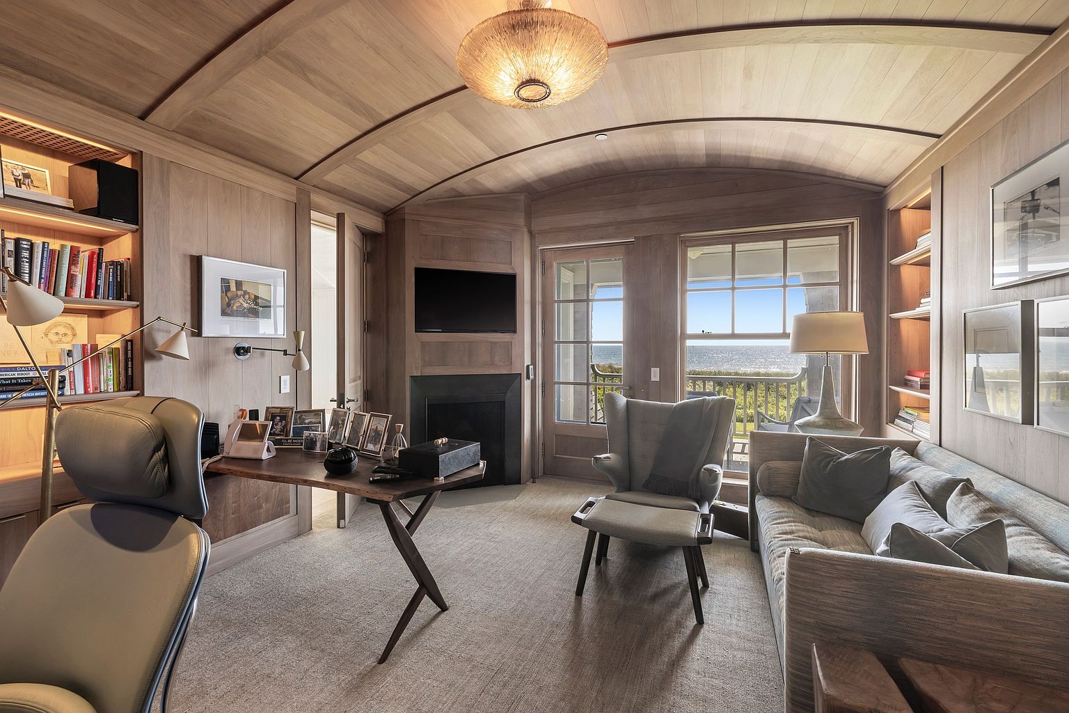 This sophisticated home office features warm, light wood paneling throughout, including a curved ceiling that adds architectural interest. The room is furnished with a sleek desk, a comfortable armchair with an ottoman, and a sofa, all oriented toward a view of the ocean through large windows and a glass-paneled door. A dark fireplace surround and built-in shelving units complete the refined, cozy aesthetic of this workspace.