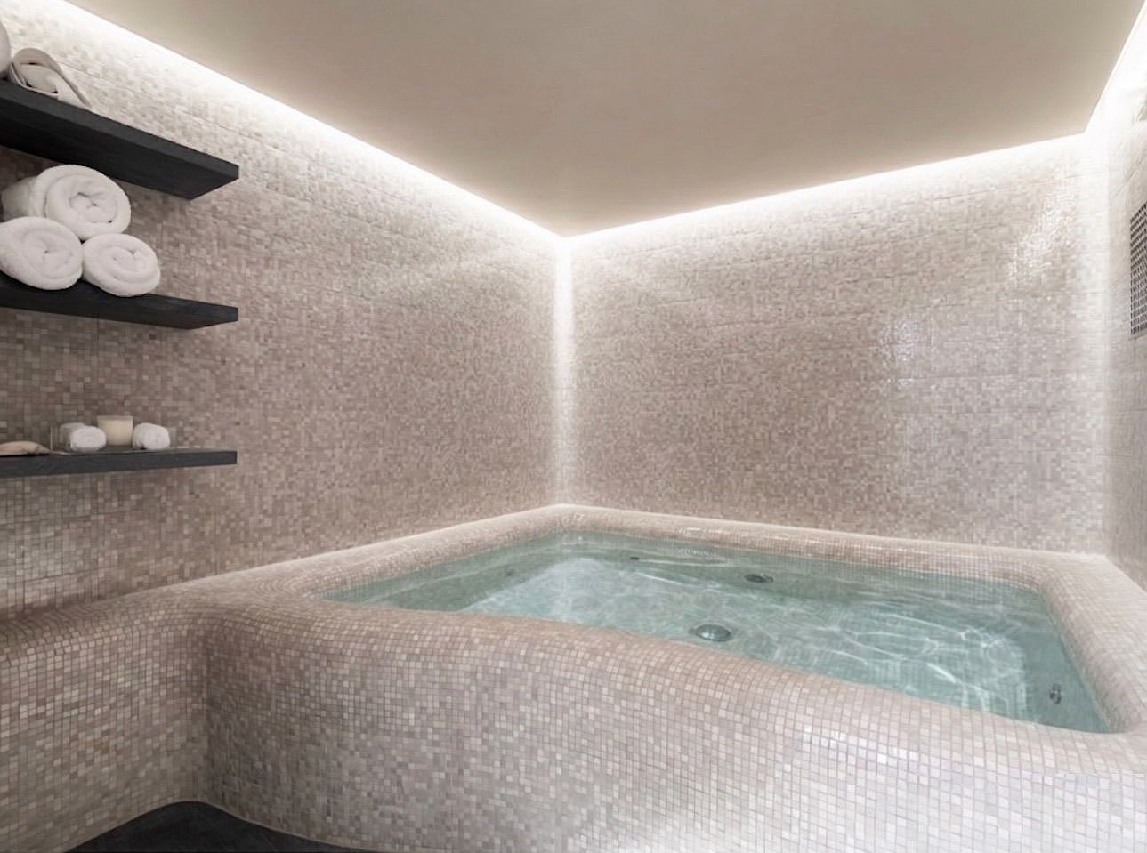 This serene spa-like bathroom features a custom-built, tiled soaking tub integrated into the corner of the room. The walls and tub are finished in a neutral, light-toned mosaic tile, complemented by recessed cove lighting that creates a soft, ambient glow. Floating shelves on the left provide organized storage for towels and spa accessories, contributing to the minimalist and tranquil aesthetic.
