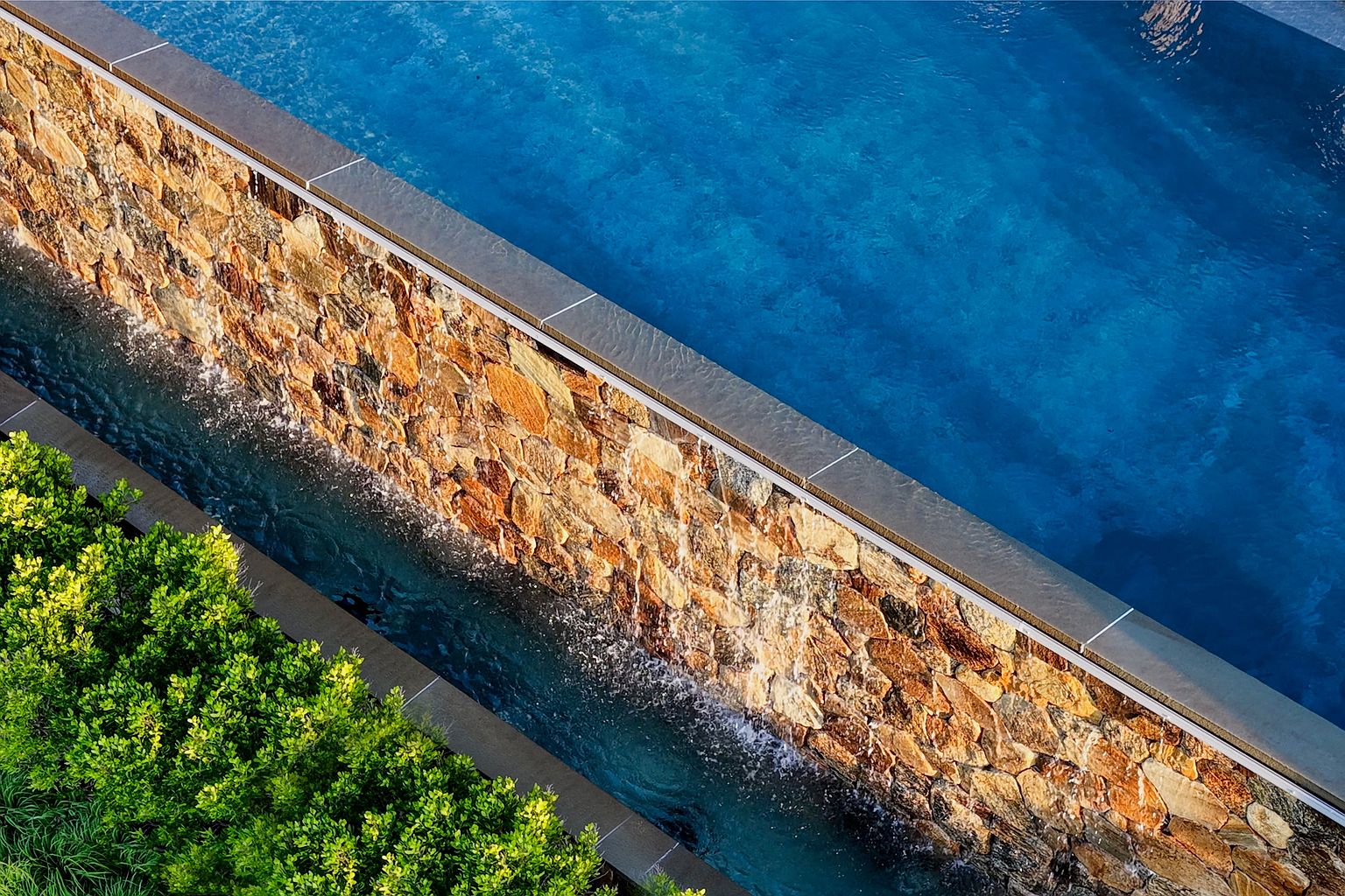 This high-angle, cinematic shot captures a luxurious infinity-edge pool featuring a textured stone retaining wall that doubles as a water feature. Water cascades down the face of the stone wall into a lower channel, creating a serene and sophisticated atmosphere. Lush green foliage lines the edge of the lower basin, providing a vibrant contrast to the deep blue water and the warm, earthy tones of the stone masonry.