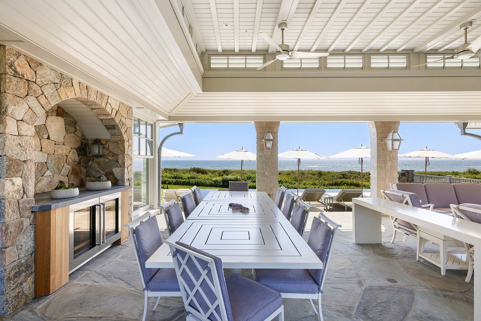 This expansive covered outdoor dining area features a stone-clad wall with a built-in beverage cooler and a large dining table set for multiple guests. The space is open to a scenic coastal view, showcasing a swimming pool, lush greenery, and the ocean beyond under a bright sky. The architectural design includes a white paneled ceiling with integrated fans and stone flooring, creating a sophisticated and airy atmosphere perfect for al fresco entertaining.