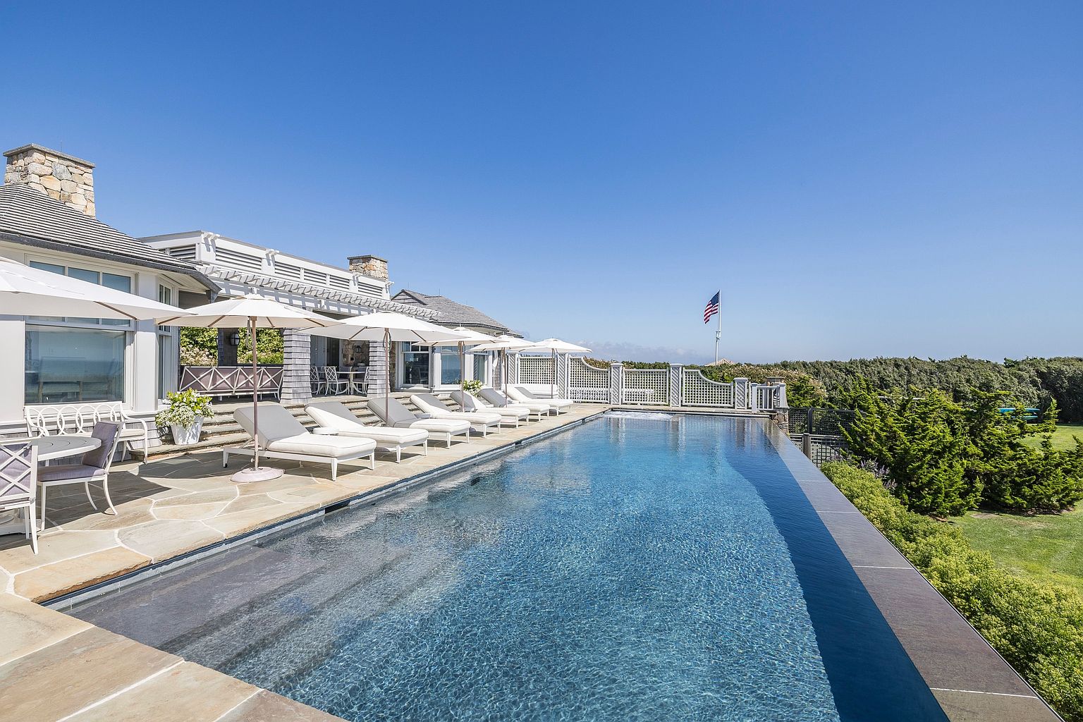 This expansive outdoor living space features a sleek infinity-edge pool that overlooks a lush, green landscape under a clear blue sky. A stone-paved patio area is furnished with multiple white lounge chairs shaded by umbrellas, creating a serene and luxurious atmosphere. The architecture of the home includes a covered porch with a pergola, blending classic coastal design with modern resort-style amenities.