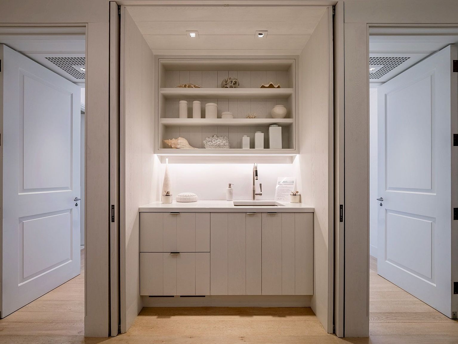 This image captures a sophisticated, built-in wet bar or butler's pantry nestled between two doorways in a bright, neutral-toned hallway. The space features light wood cabinetry with integrated shelving above, illuminated by soft under-cabinet and recessed ceiling lighting. The clean, minimalist aesthetic is accented by decorative coastal-inspired objects on the shelves, creating a serene and functional transition area.